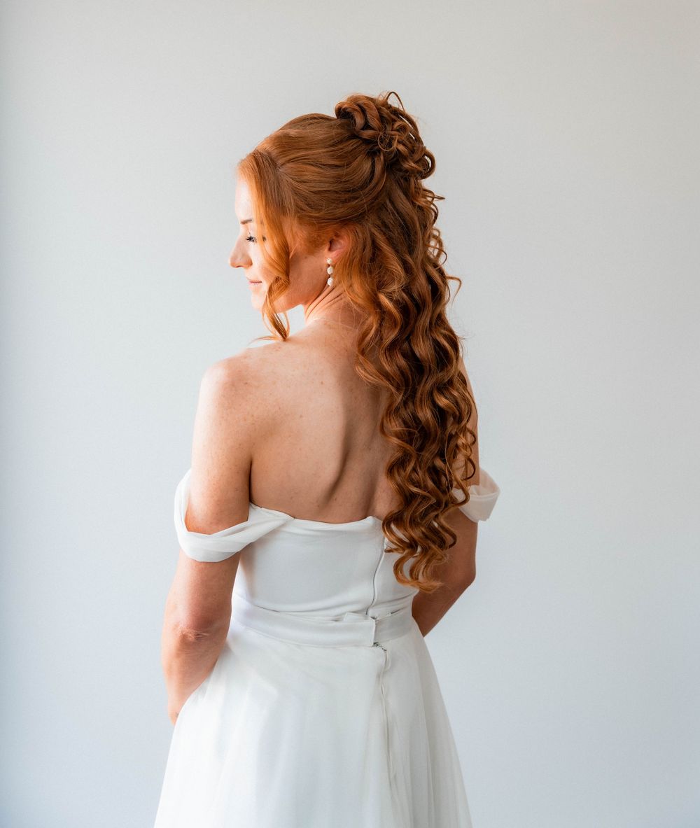 Bride with long curly red hair in half-up style wearing white off-shoulder wedding dress from behind.