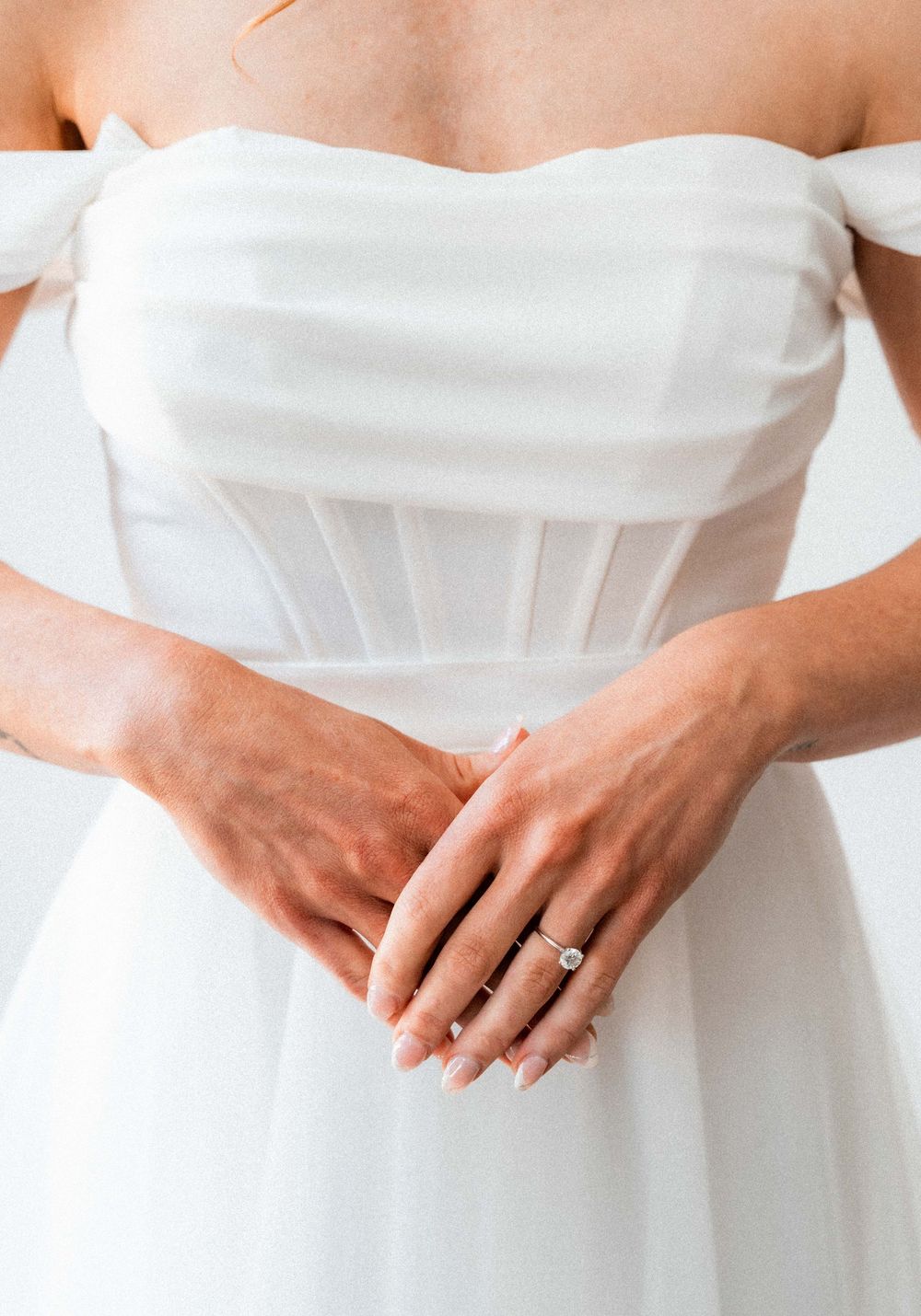Bride in white off-shoulder corset wedding gown clasping hands, showcasing elegant diamond engagement ring.