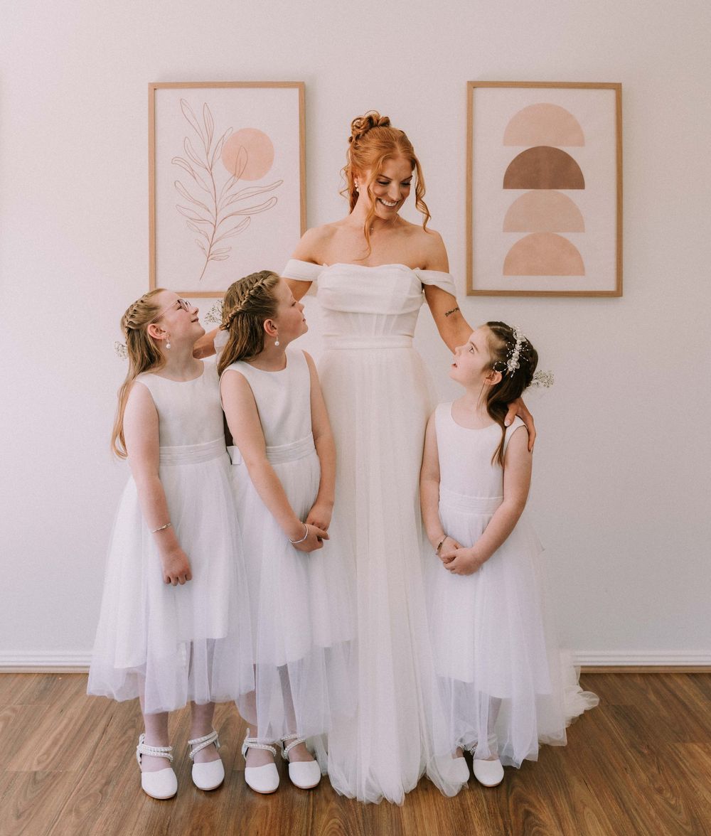 Bride in white off-shoulder gown smiling with three flower girls in white tulle dresses indoors.