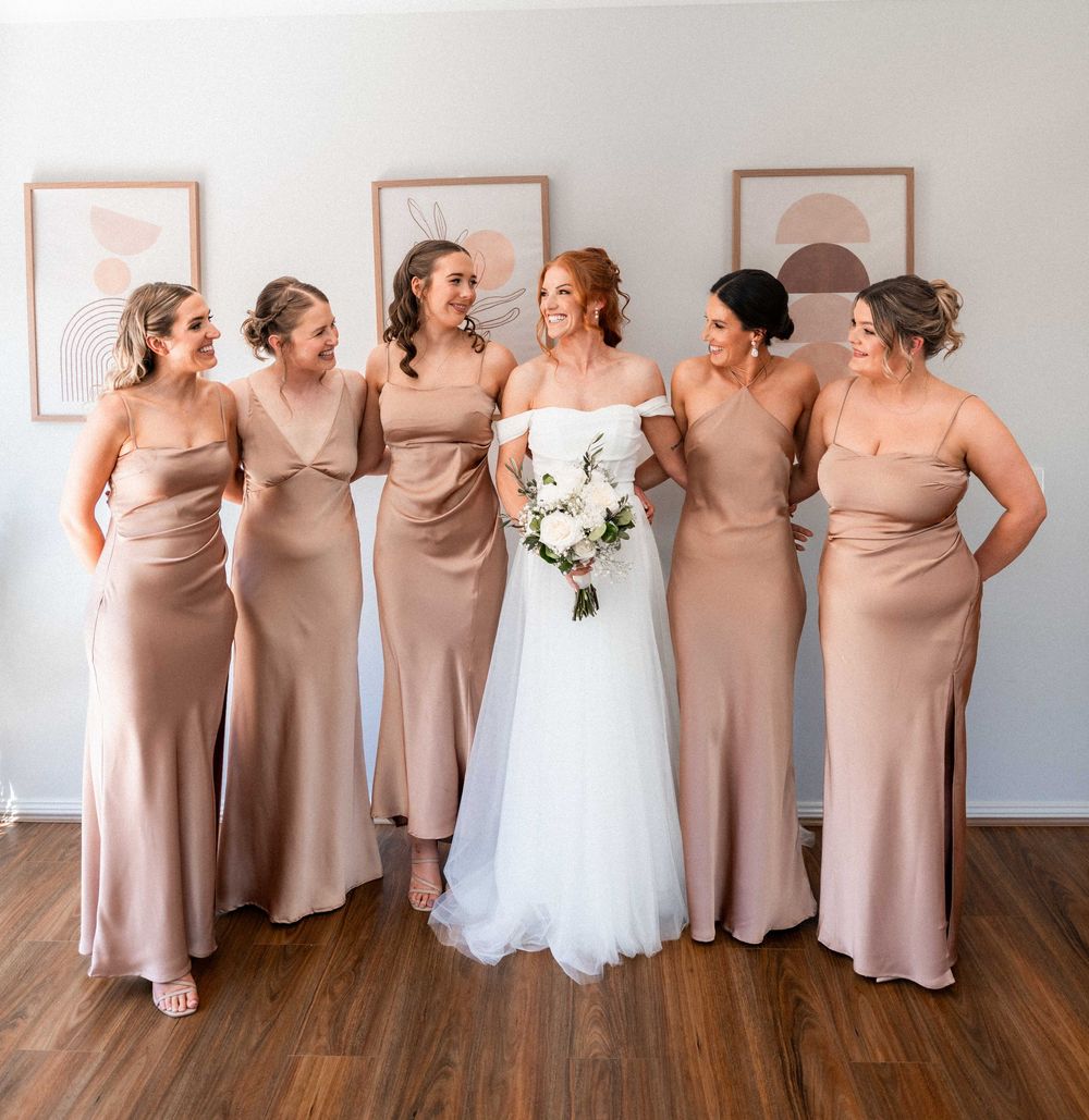 Bride in white gown holds bouquet surrounded by four bridesmaids in champagne satin dresses, smiling together indoors.