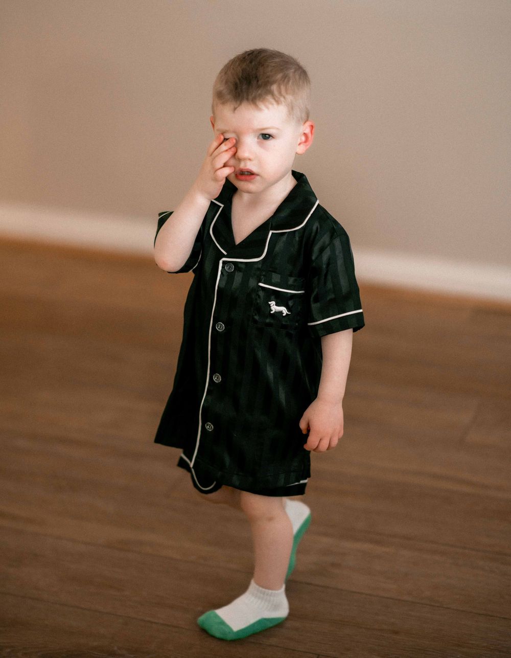 Toddler boy in dark green velvet pajama set with white piping, rubbing his eye while standing on hardwood floor.