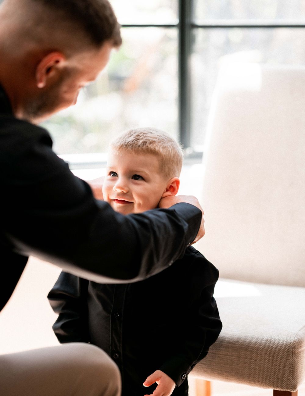 Father helping young toddler boy put on a black jacket while sitting near a bright window indoors.