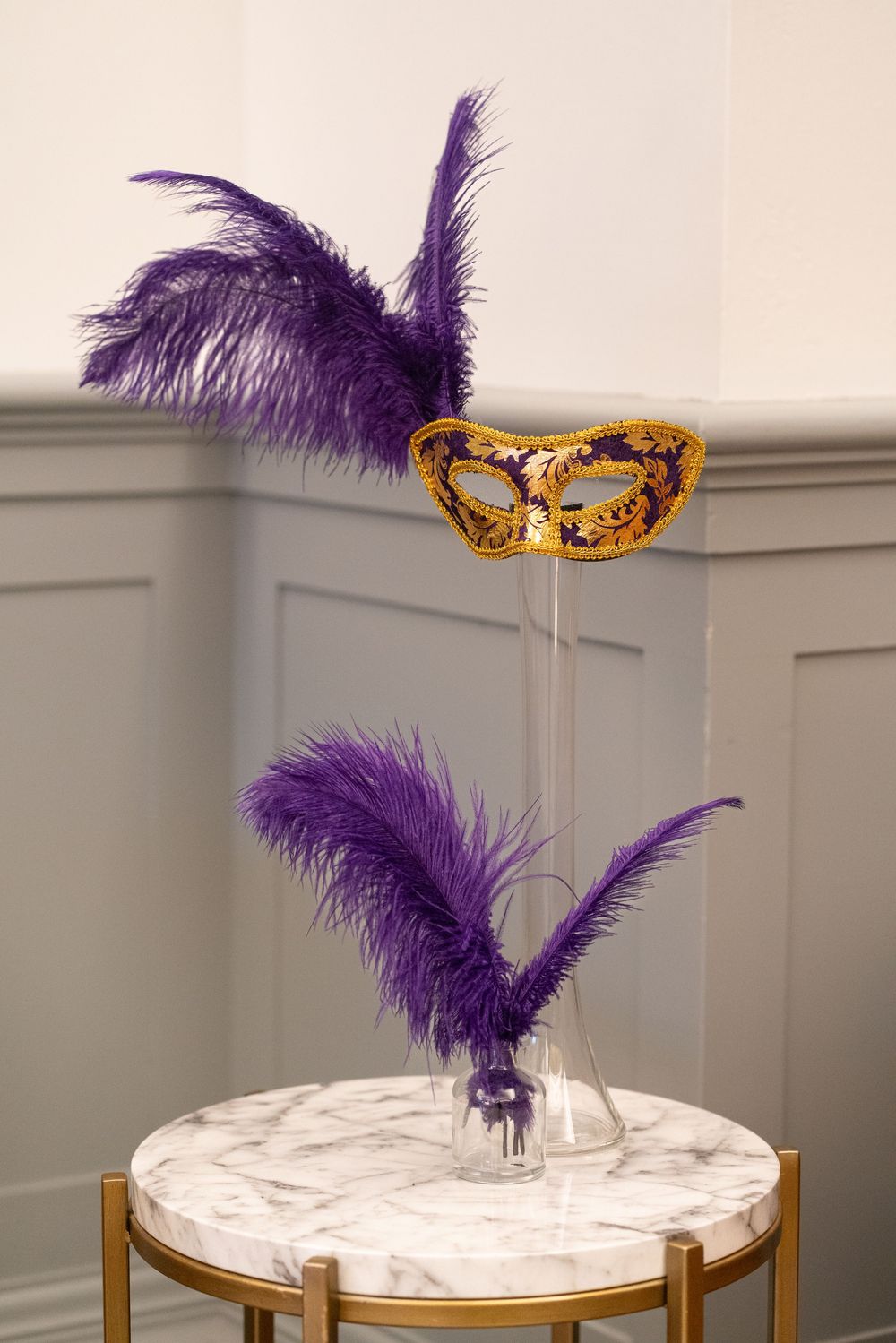 Gold and purple Venetian masquerade mask with feathers displayed on a marble table against gray wainscoting wall.