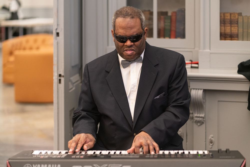 Man in suit and sunglasses playing a Yamaha keyboard piano in an elegant room.