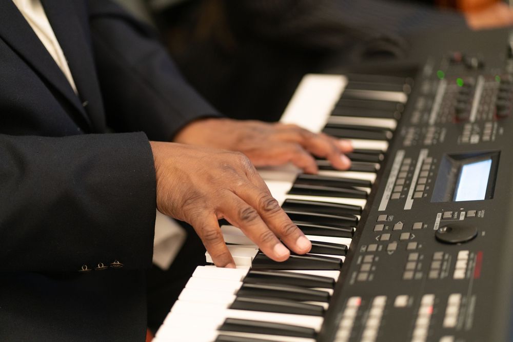 A musician plays a digital keyboard synthesizer, with hands visible on black and white keys, wearing a dark suit jacket.