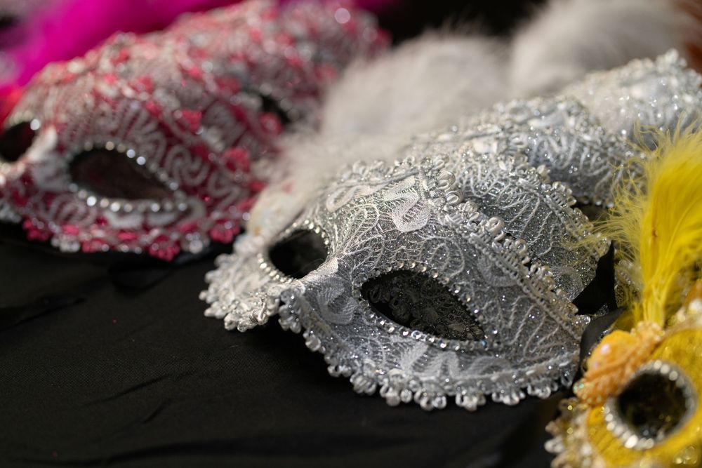 Ornate silver and pink masquerade masks with jewels and feathers displayed on a black surface.