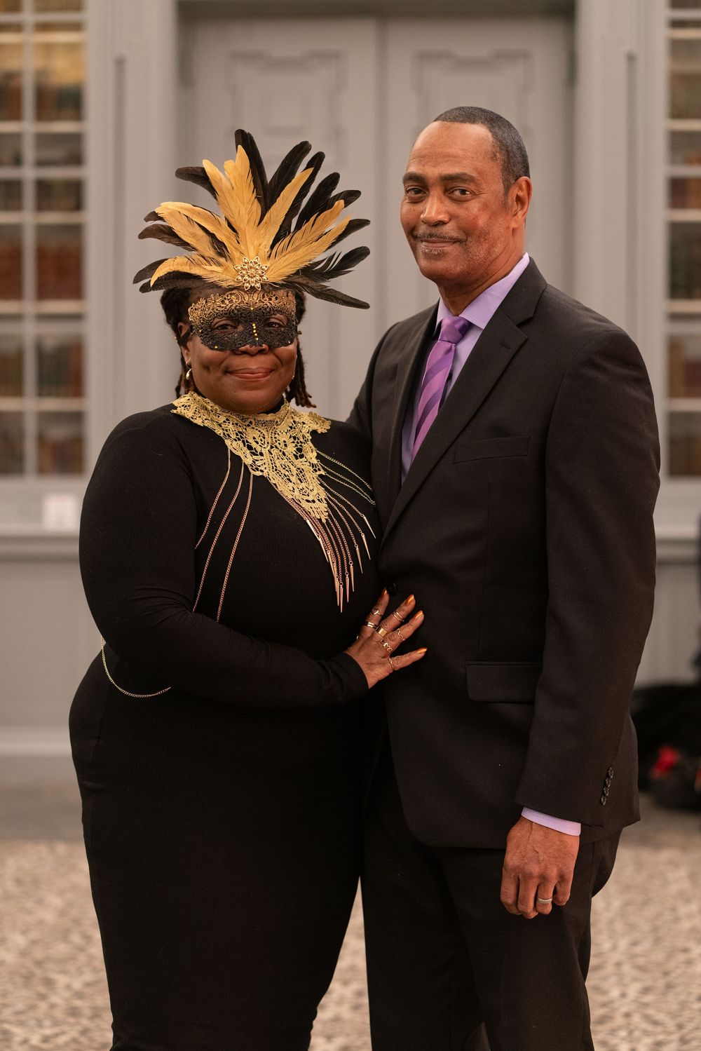 Couple posing at masquerade event, woman wearing gold feathered mask and black outfit, man in dark suit with purple tie.
