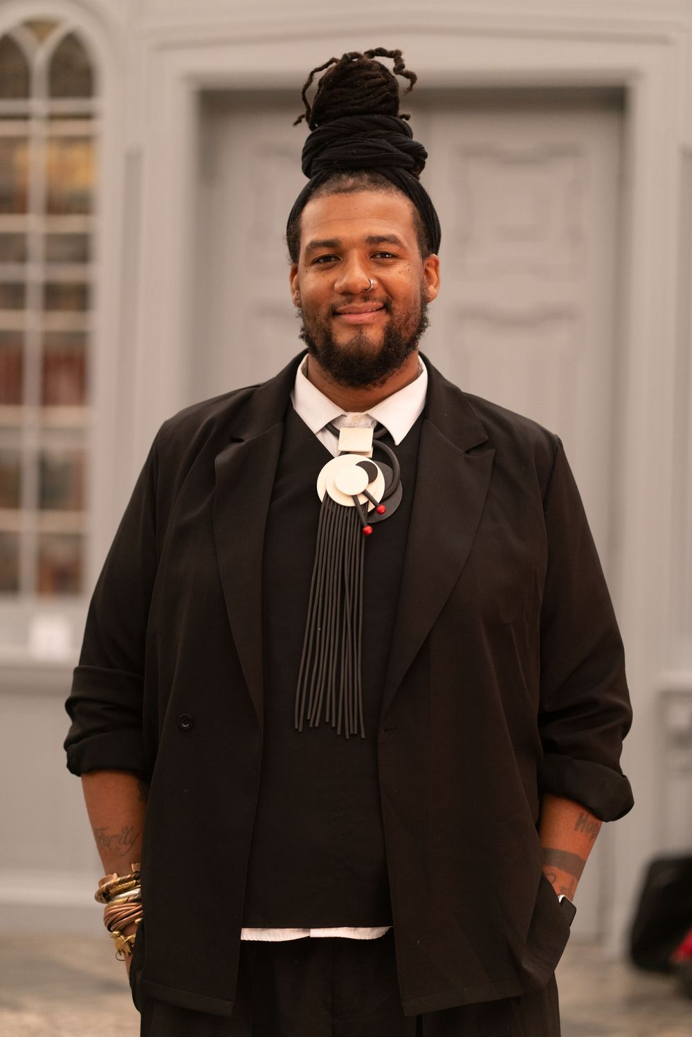Smiling man with dreadlocks in a black blazer and unique bolo tie accessory poses outdoors.
