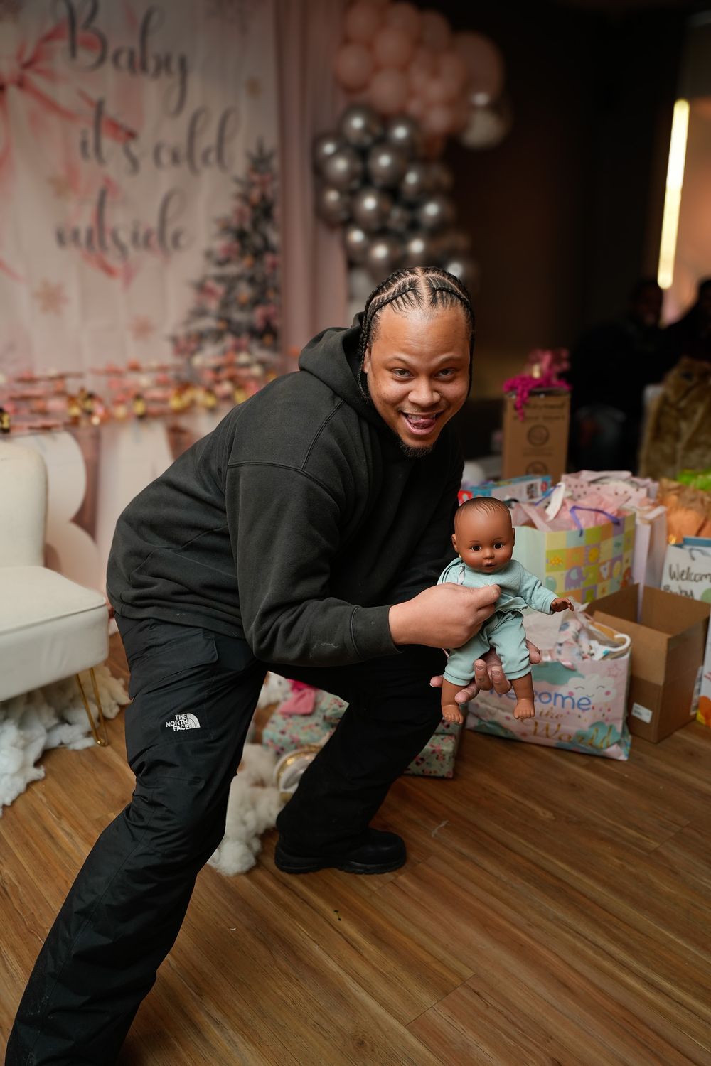 Man in black North Face hoodie posing with a baby doll at a winter-themed baby shower.