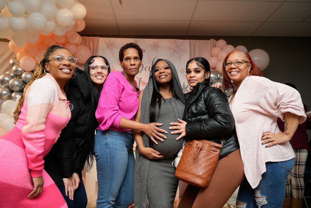Group of six women posing together at a pink and white baby shower, one visibly pregnant.