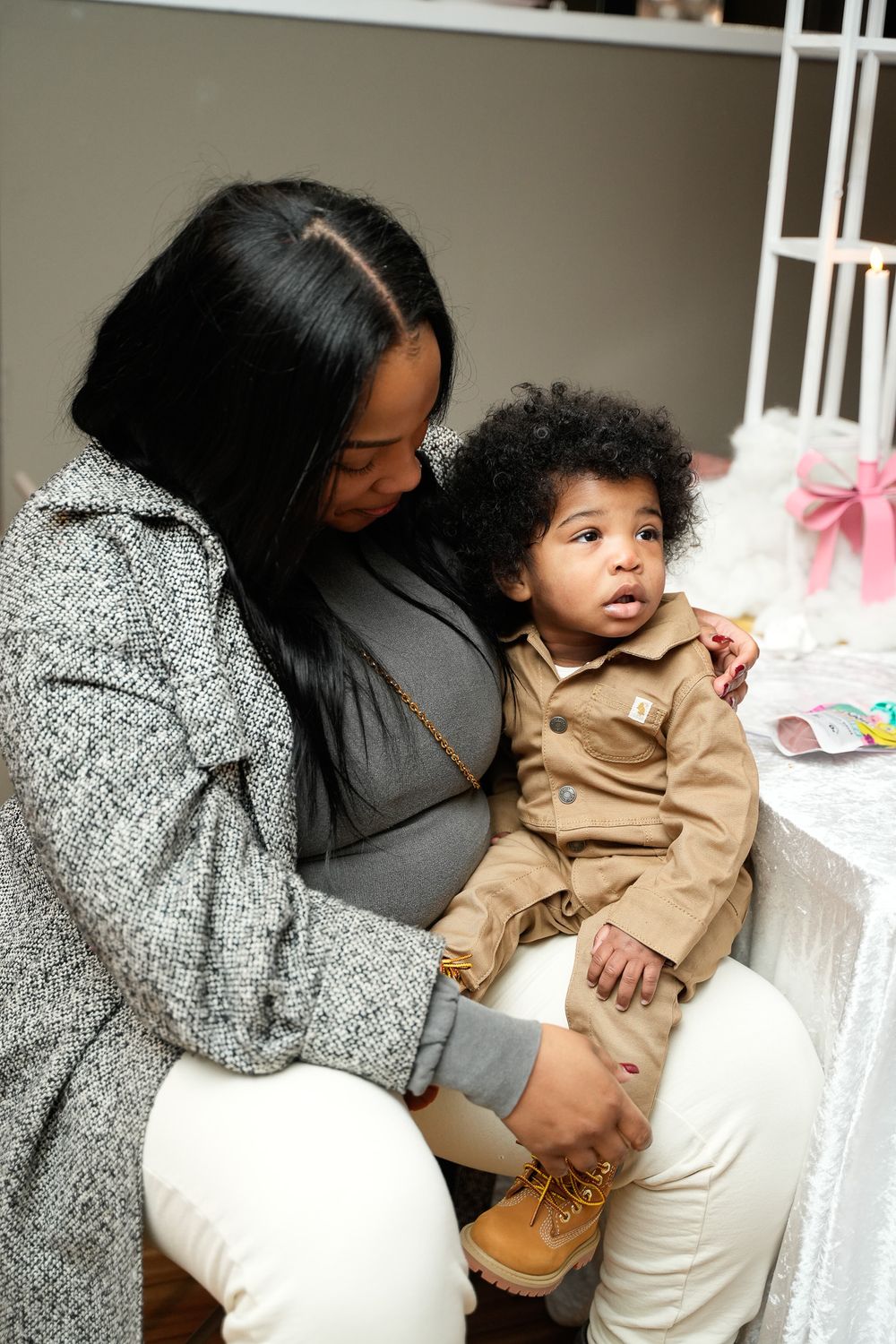 A pregnant woman in a gray coat lovingly holds a curly-haired toddler in a tan jacket at a baby shower.