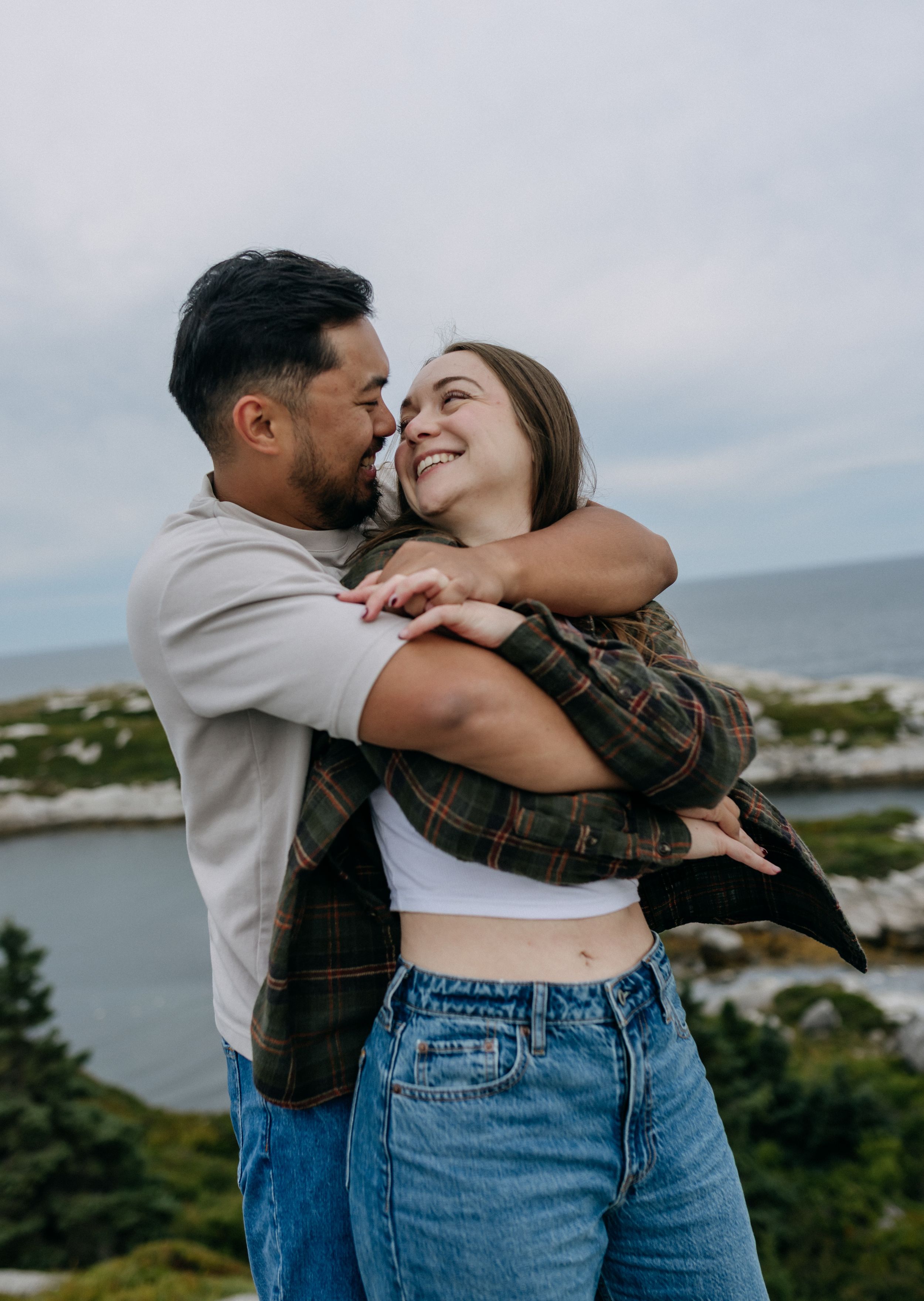 A happy couple embraces and smiles at each other outdoors near a scenic coastal landscape on a cloudy day.