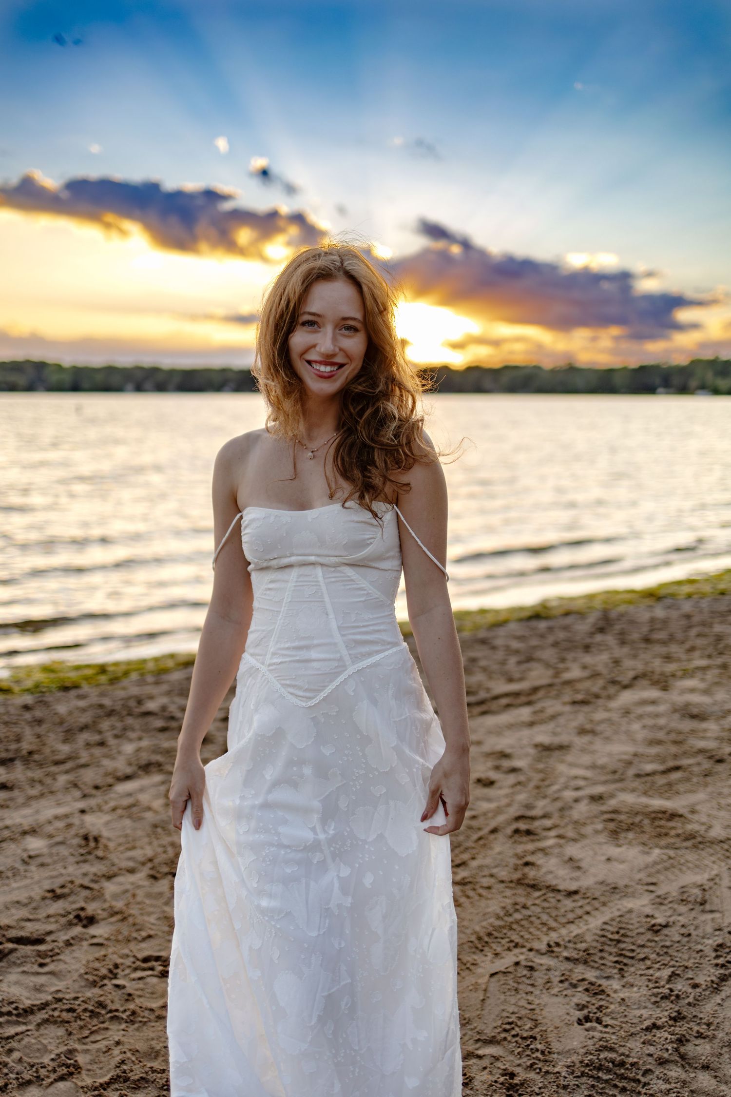 Smiling redhead woman in white strapless dress standing on sandy beach at golden sunset with dramatic clouds.