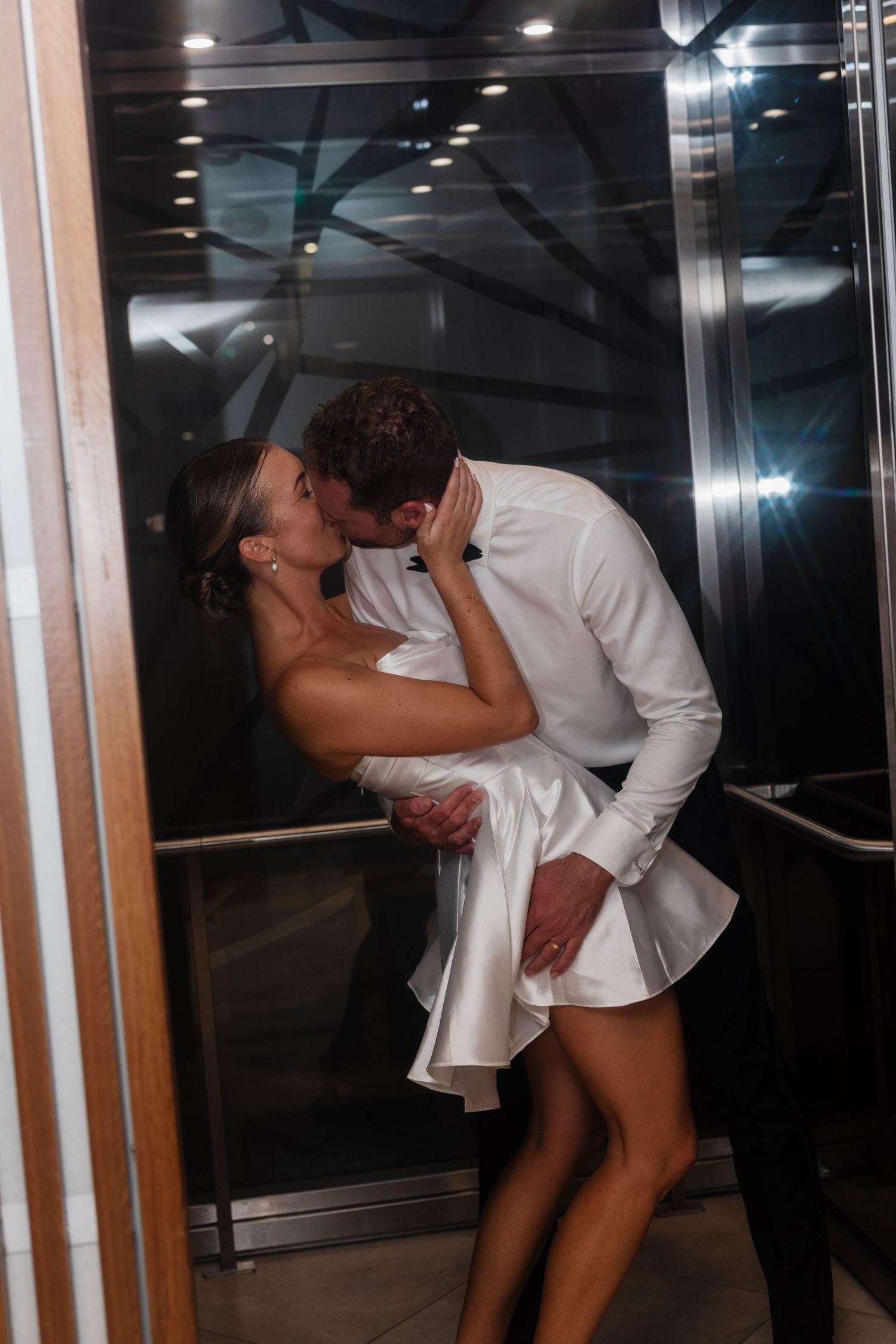 Couple sharing a romantic kiss in an elevator, bride in white mini dress and groom in white shirt.