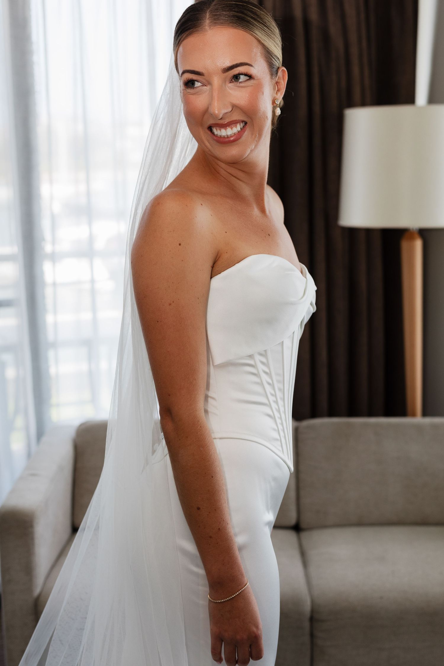 Smiling bride in strapless white corset gown and veil poses elegantly in a bright hotel room.