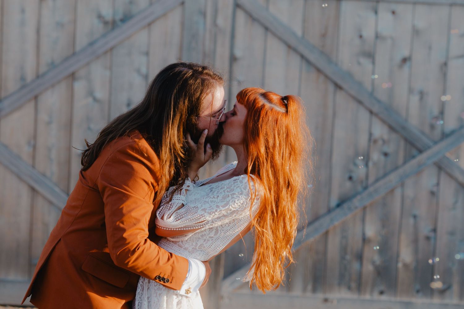 Couple sharing a romantic kiss outdoors, woman with long red hair in white dress, man in orange blazer, rustic barn backdrop.
