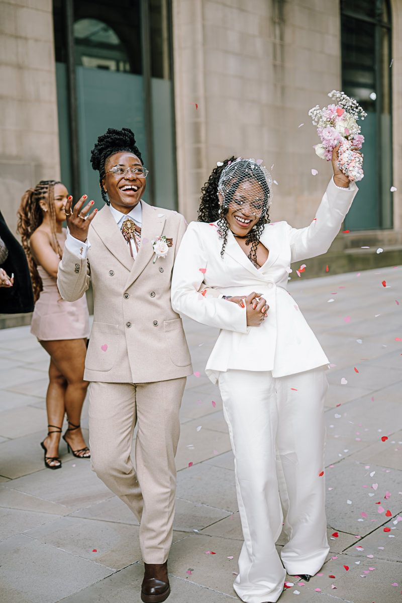 Two brides in elegant suits celebrate outside with confetti, one in white holding a bouquet, as guests cheer behind them.