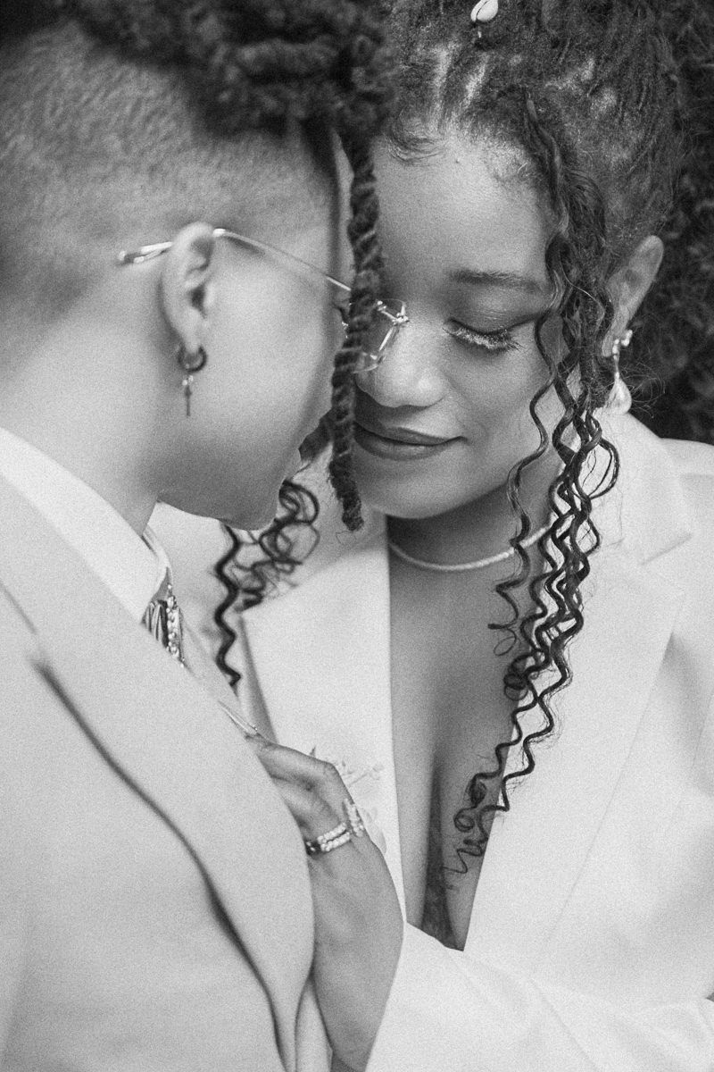 Black and white photo of two women in white blazers sharing an intimate moment, with curly hair and elegant jewelry.
