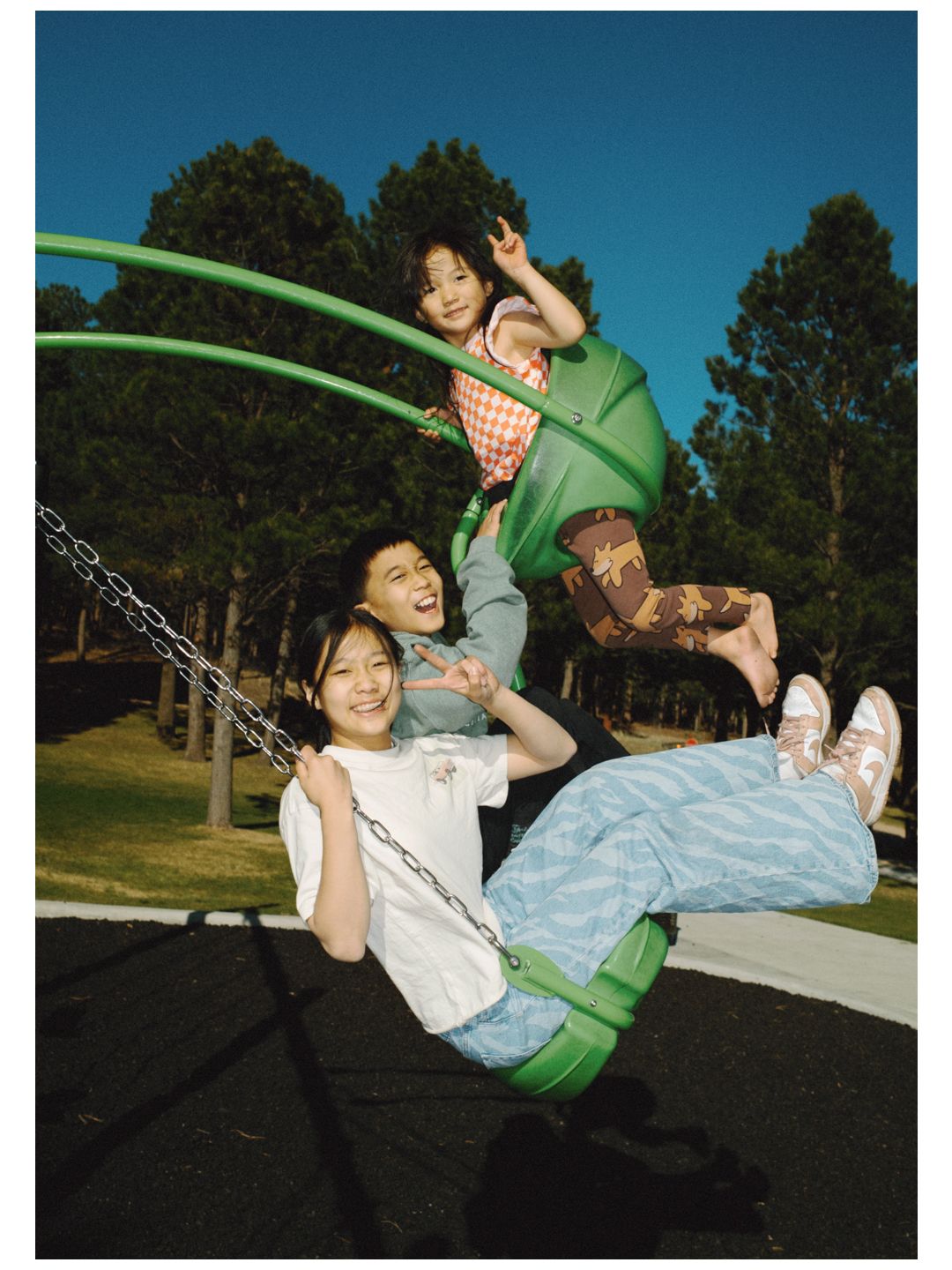 Two children and a woman laughing on a green swing set in a sunny park surrounded by tall trees.