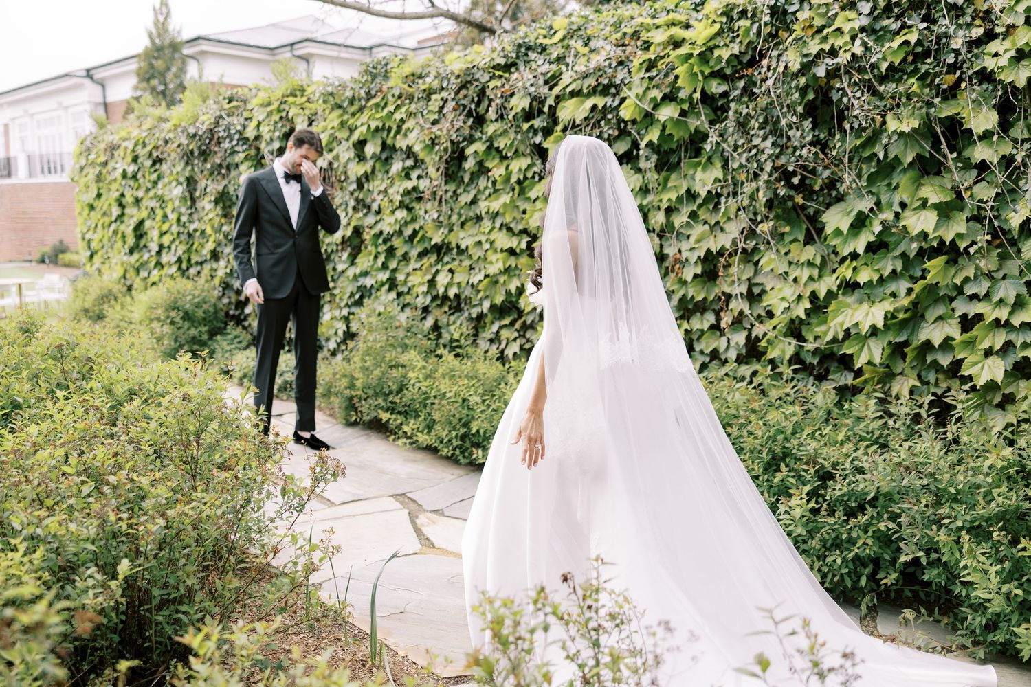 Bride in white gown and veil with groom in black suit posing in lush garden with ivy-covered wall.