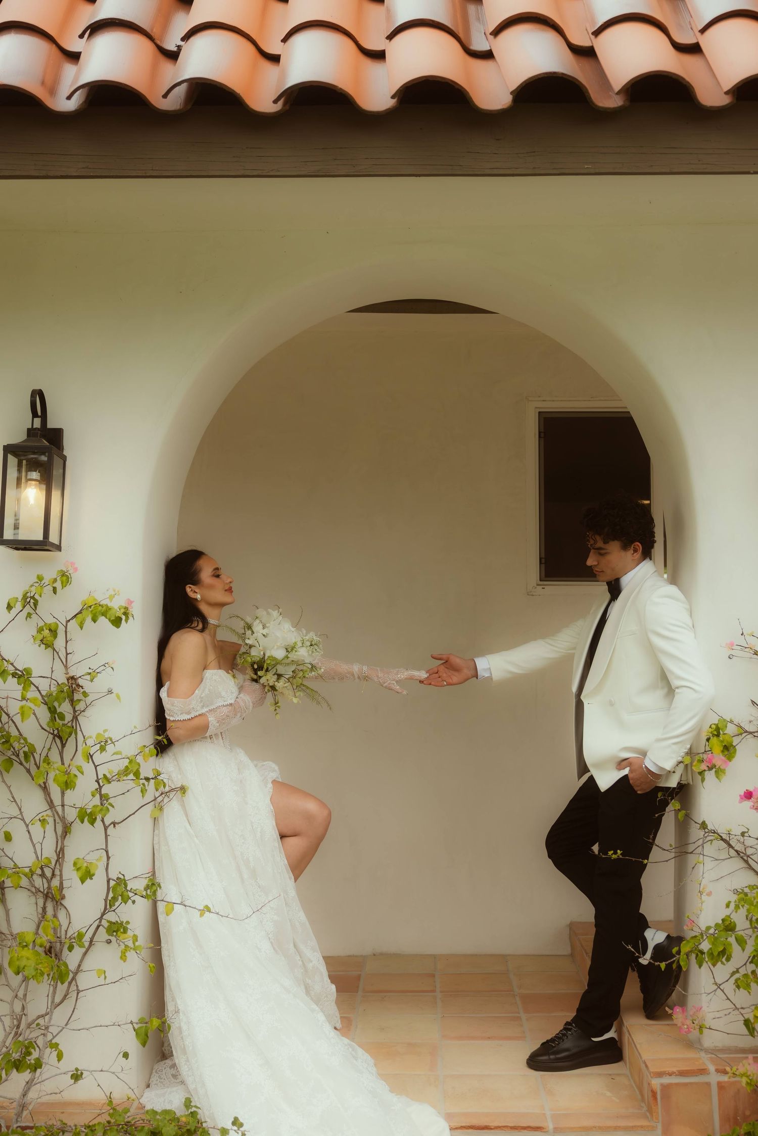 Bride in white gown holds groom's hand under arched doorway of Spanish-style home with terracotta roof.