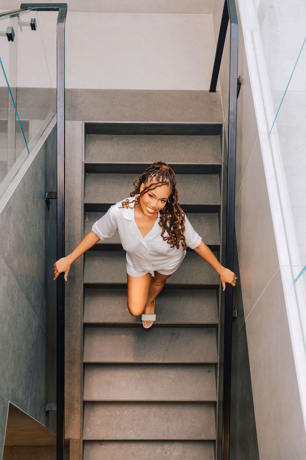 Smiling young woman with braids descending modern gray stairs in a white shirt, viewed from above.