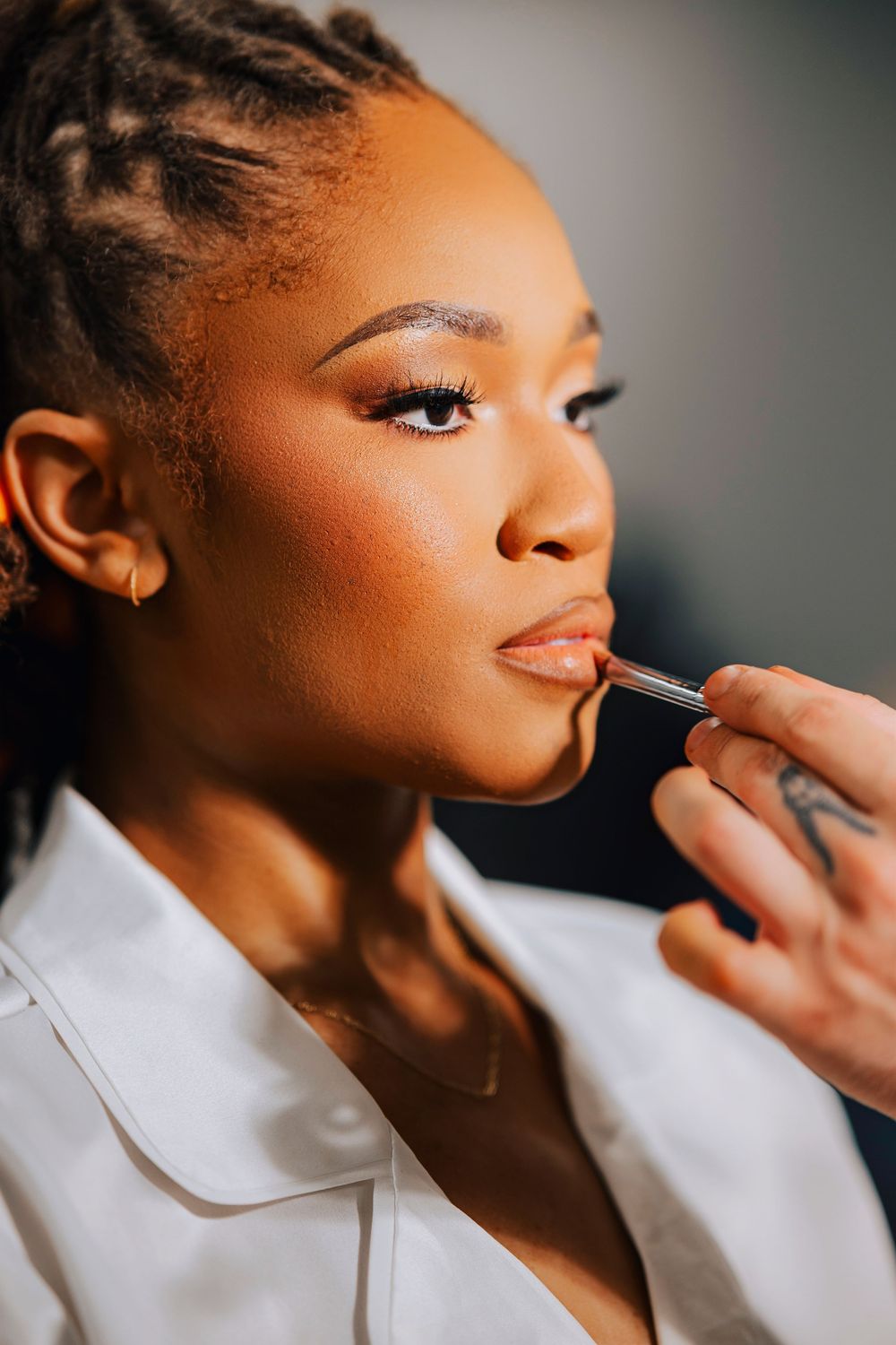 A makeup artist applies nude lip liner to a young Black woman with braids and glam eye makeup in a white top.