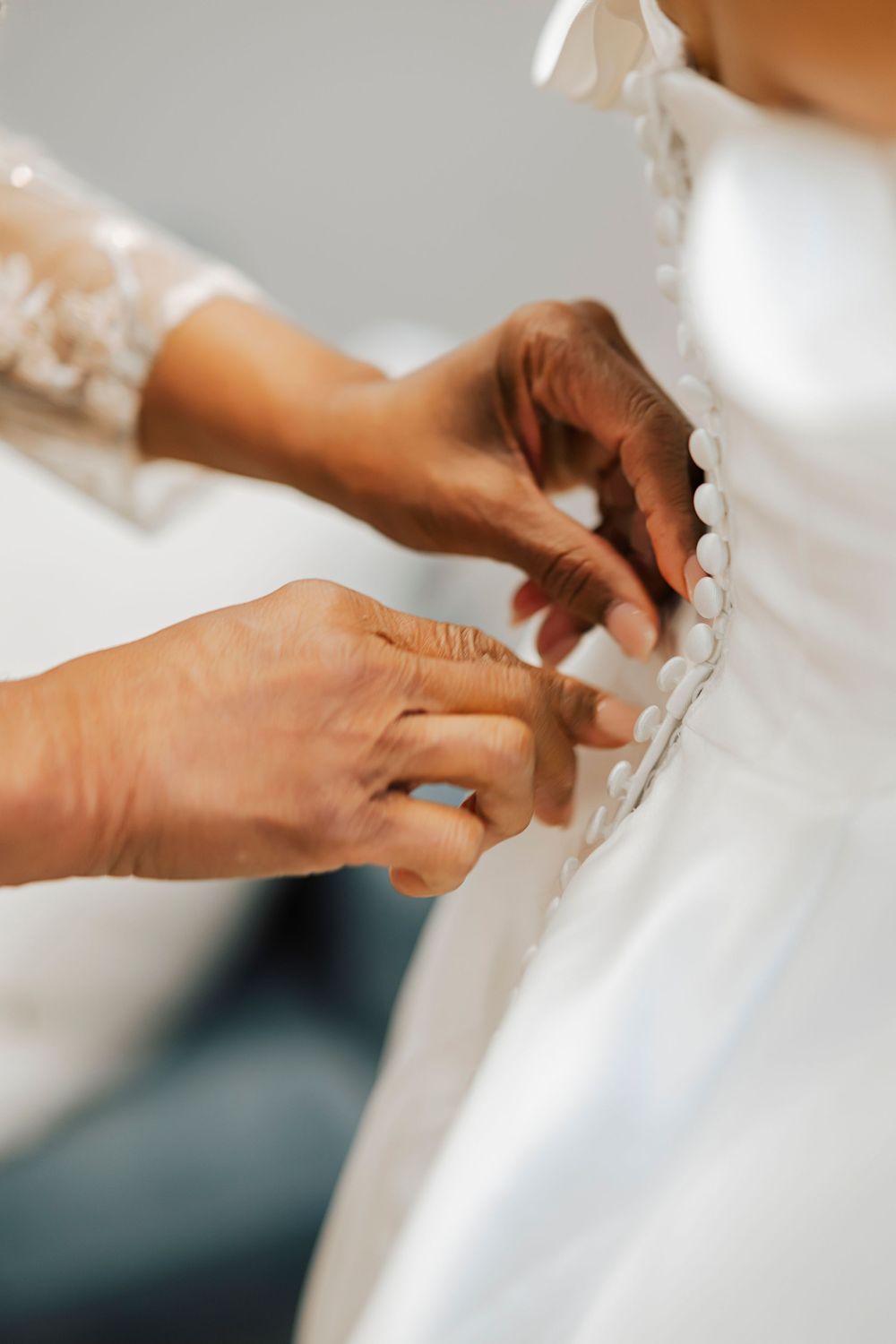 Hands carefully fastening small buttons on the back of a white wedding dress.