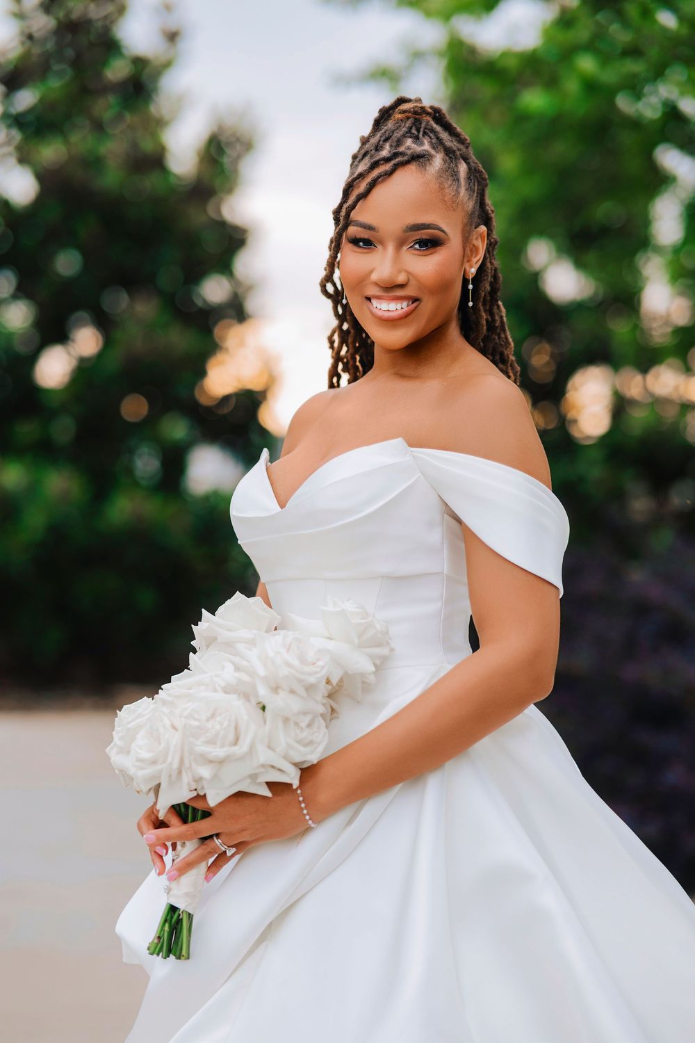 Smiling bride in off-shoulder white ball gown holding white rose bouquet, with braided updo, outdoors.