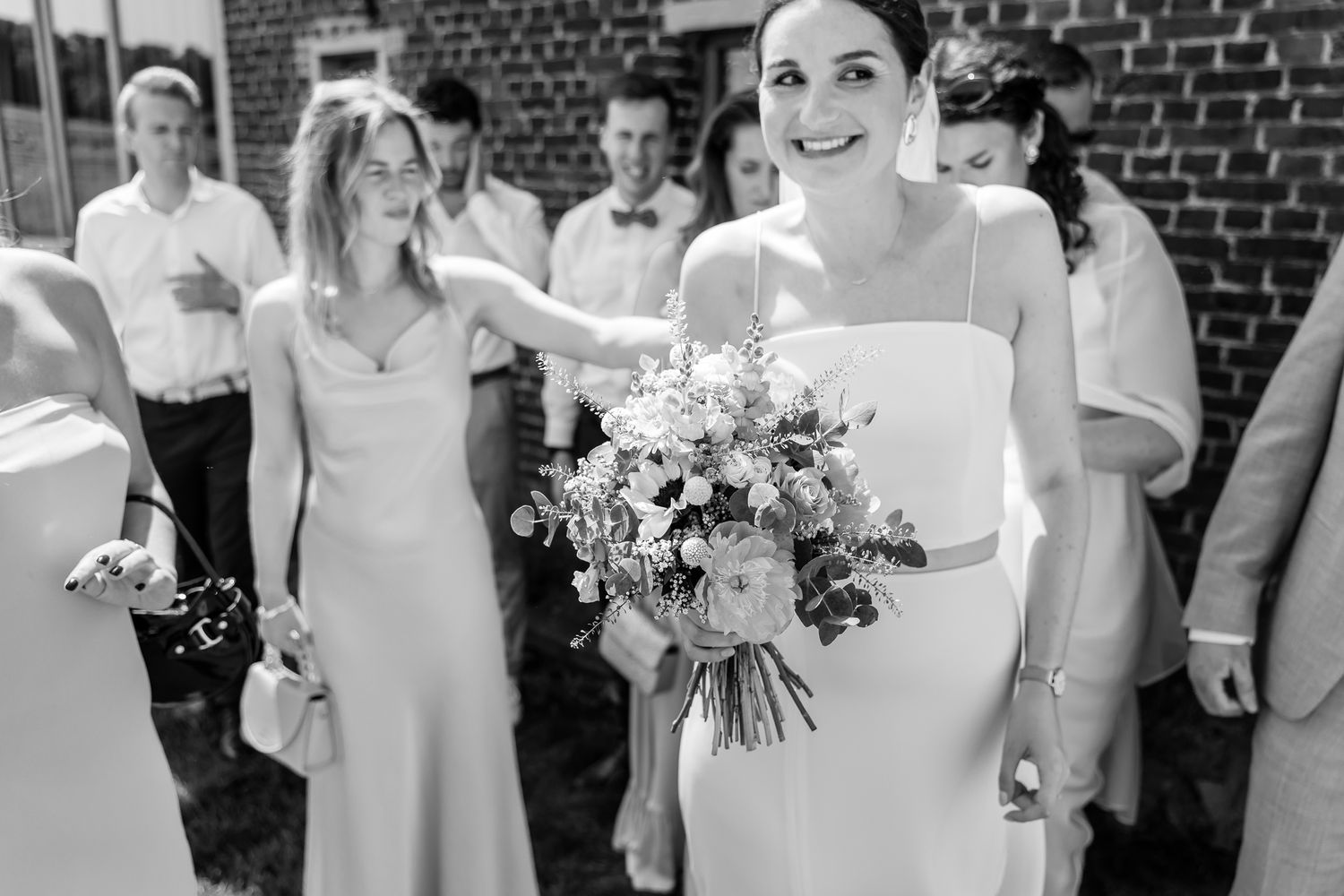 Bride in white gown holds floral bouquet surrounded by wedding guests in black and white photo.