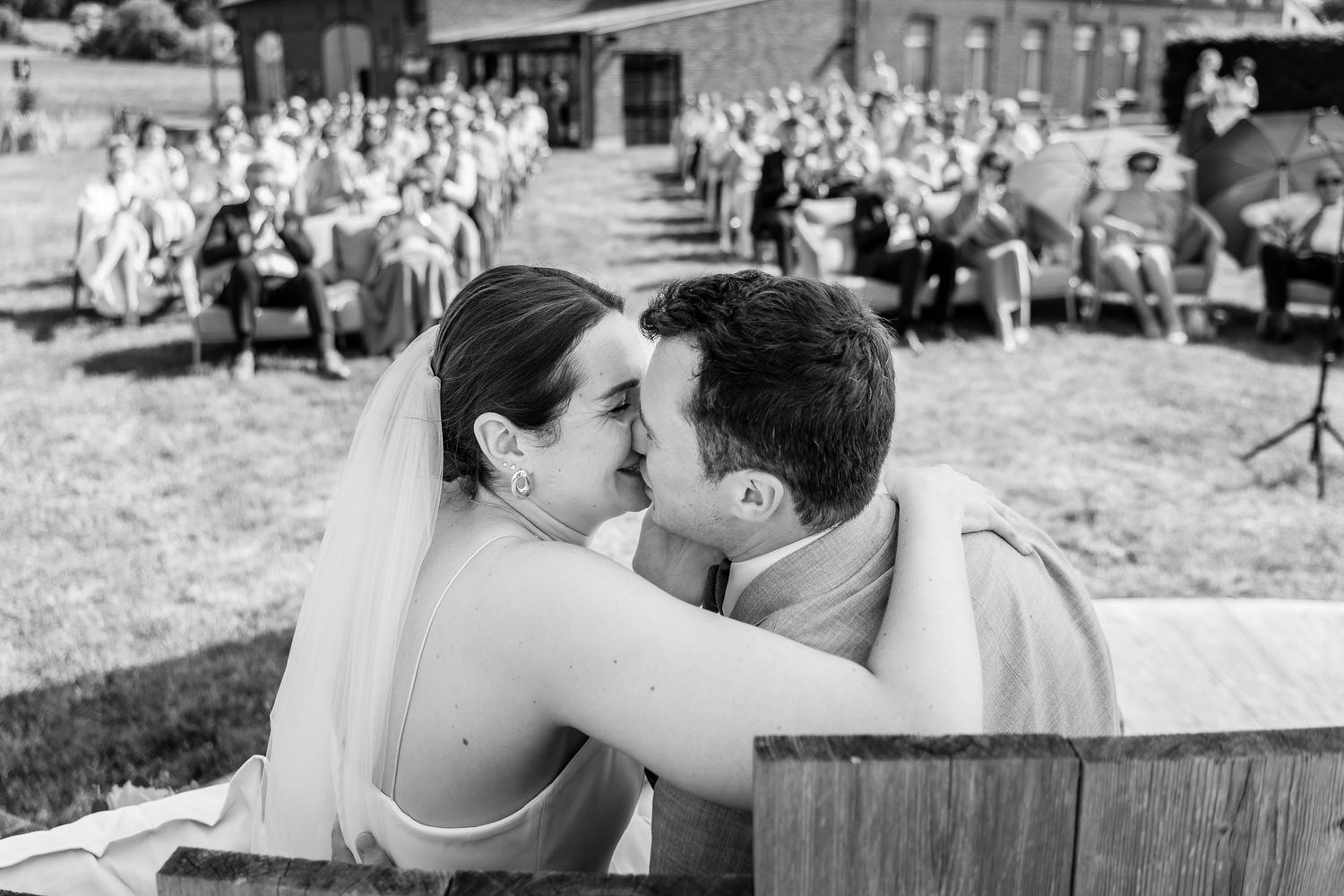 Black and white photo of bride and groom sharing a kiss at outdoor wedding ceremony with guests seated in background.