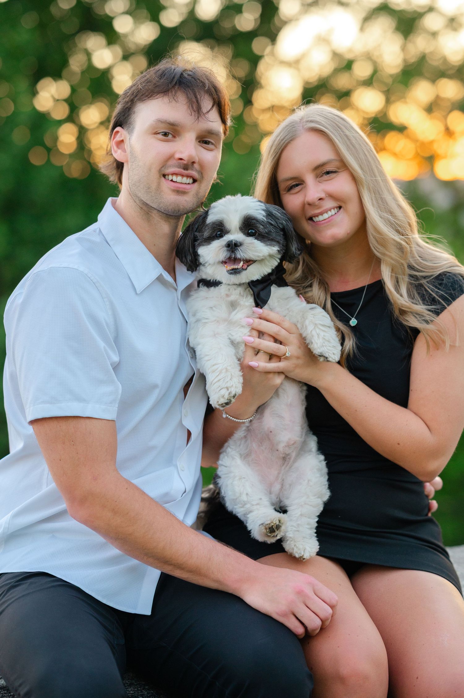 Couple holding hands on green hill with dog, overlooking scenic lake and trees at sunset after their proposal near Rockwood Hall.