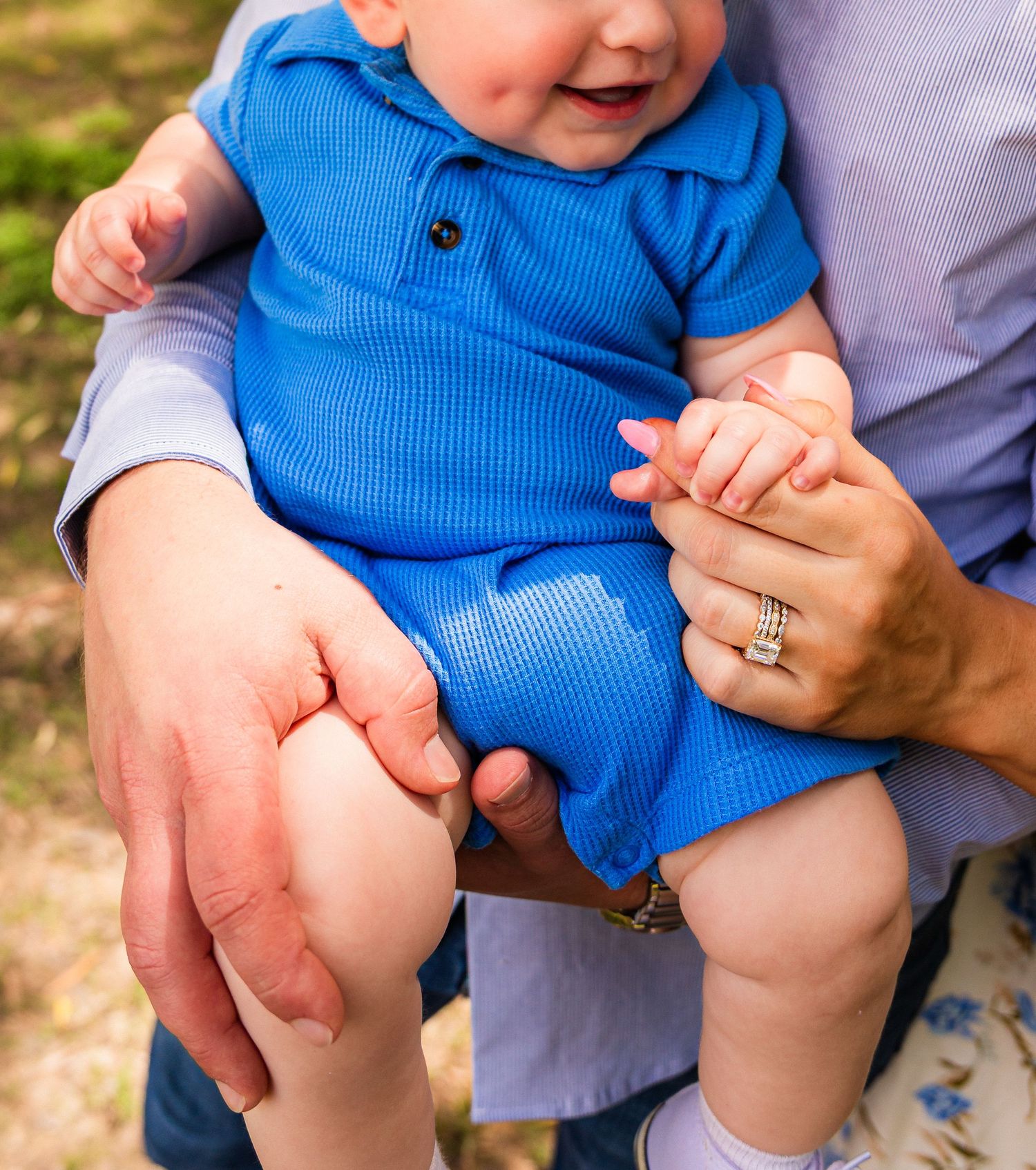 A happy baby in a blue ribbed romper being held by parents outdoors, with mom's ring visible.