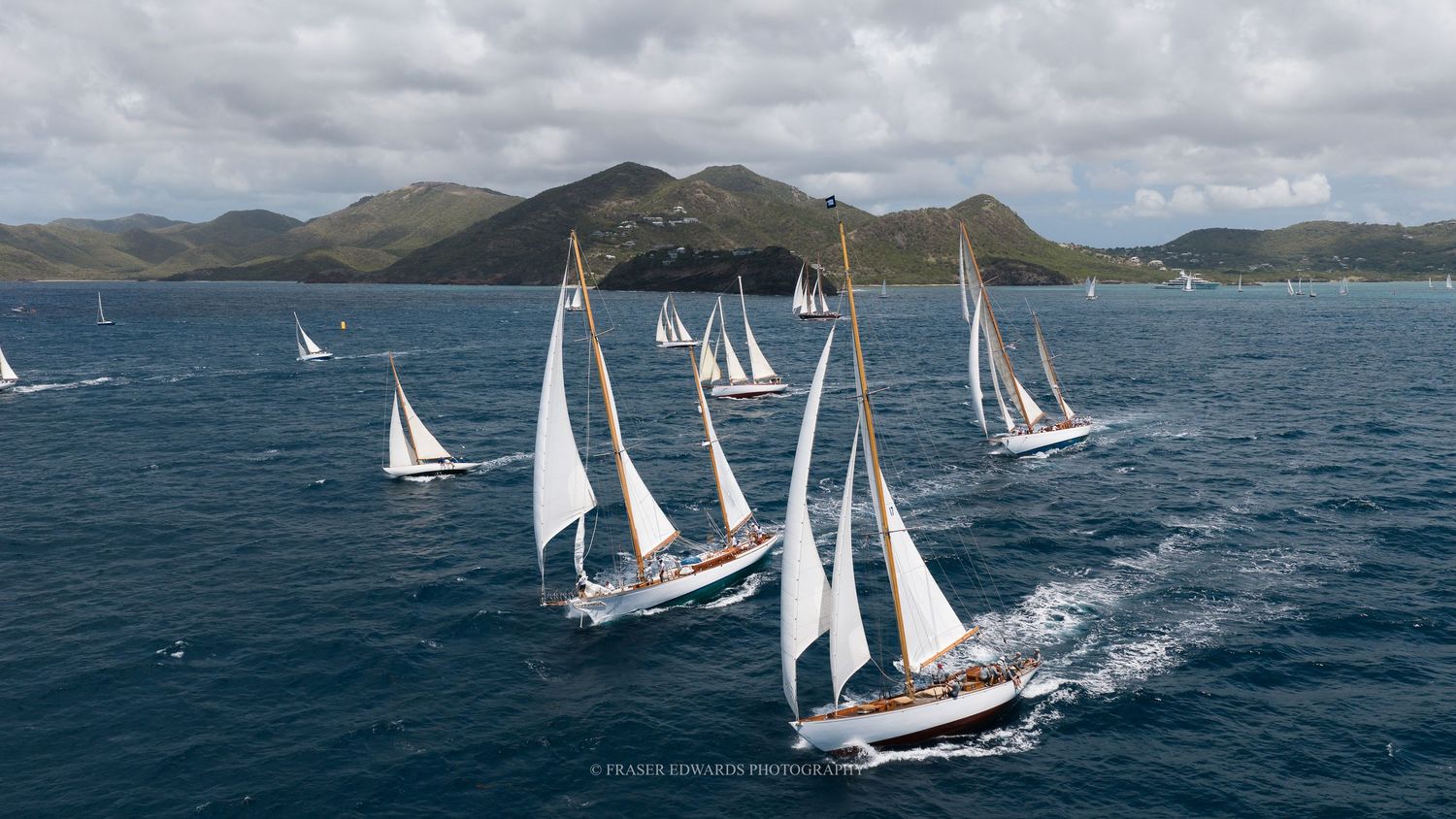 Fleet of classic sailing yachts racing on turquoise Caribbean waters with lush green hills in the background.