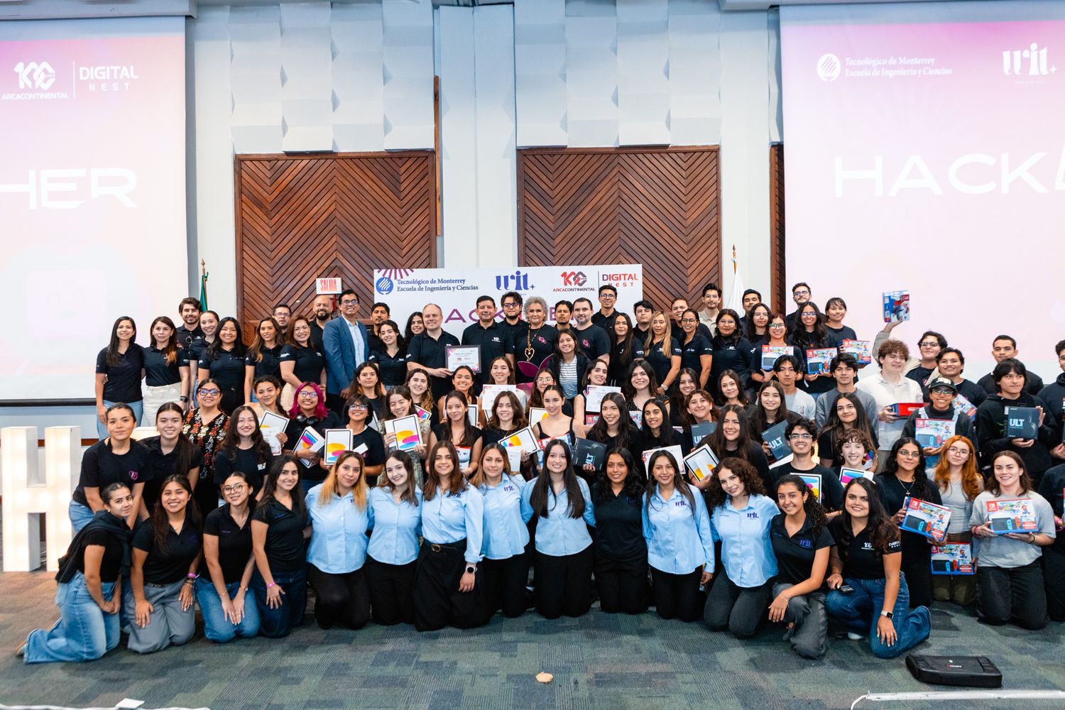 Large group of students and professionals pose together at a hackathon event in a modern conference venue.
