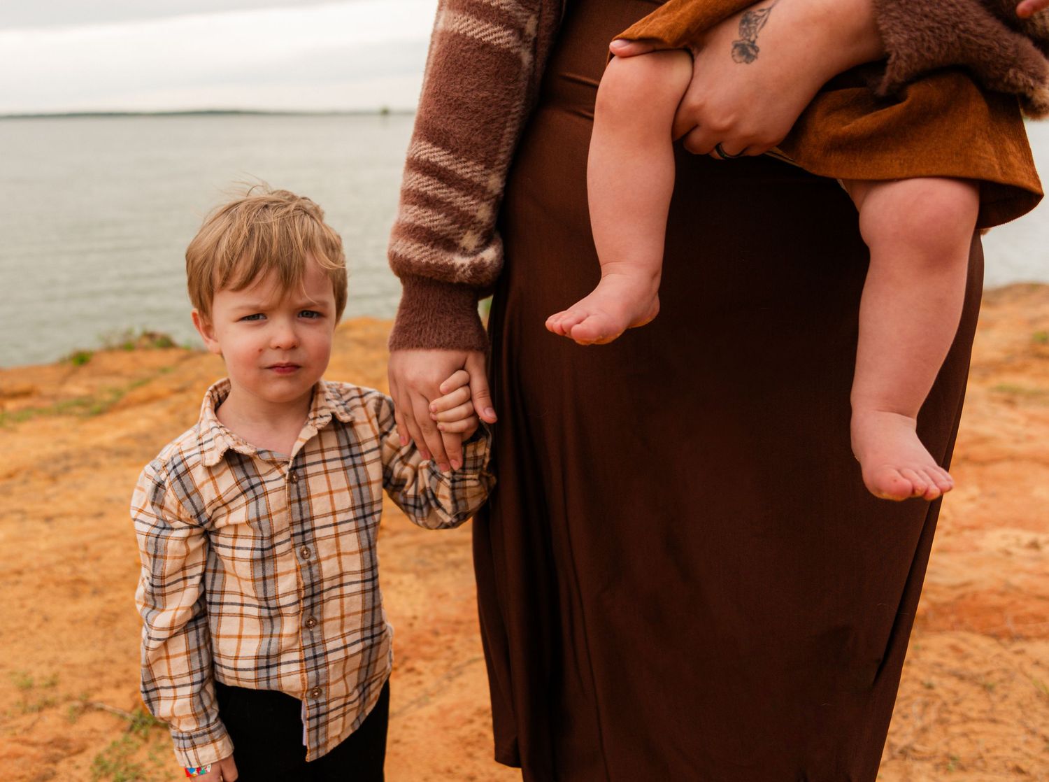 A young boy holds hands with a woman in a brown dress holding a baby near a coastal cliffside.