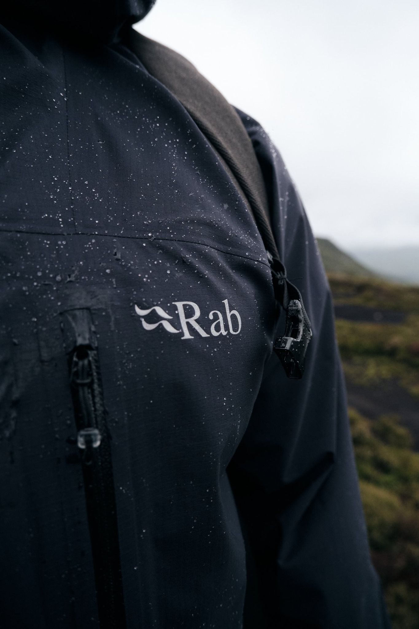 Close-up of black Rab waterproof jacket with logo, covered in rain droplets outdoors.