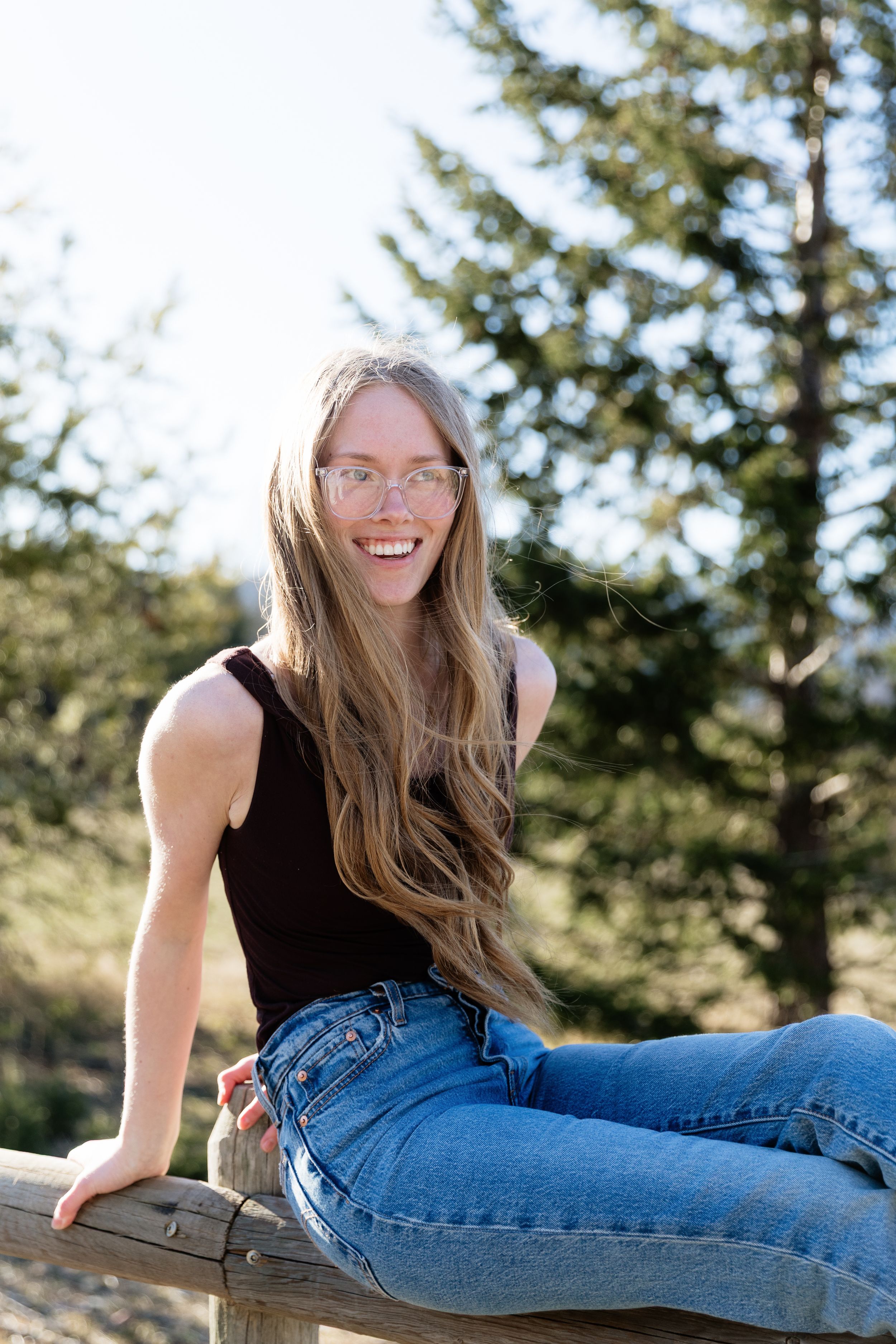 Young woman with long blonde hair and glasses smiling outdoors, wearing black tank top and blue jeans on wooden fence.