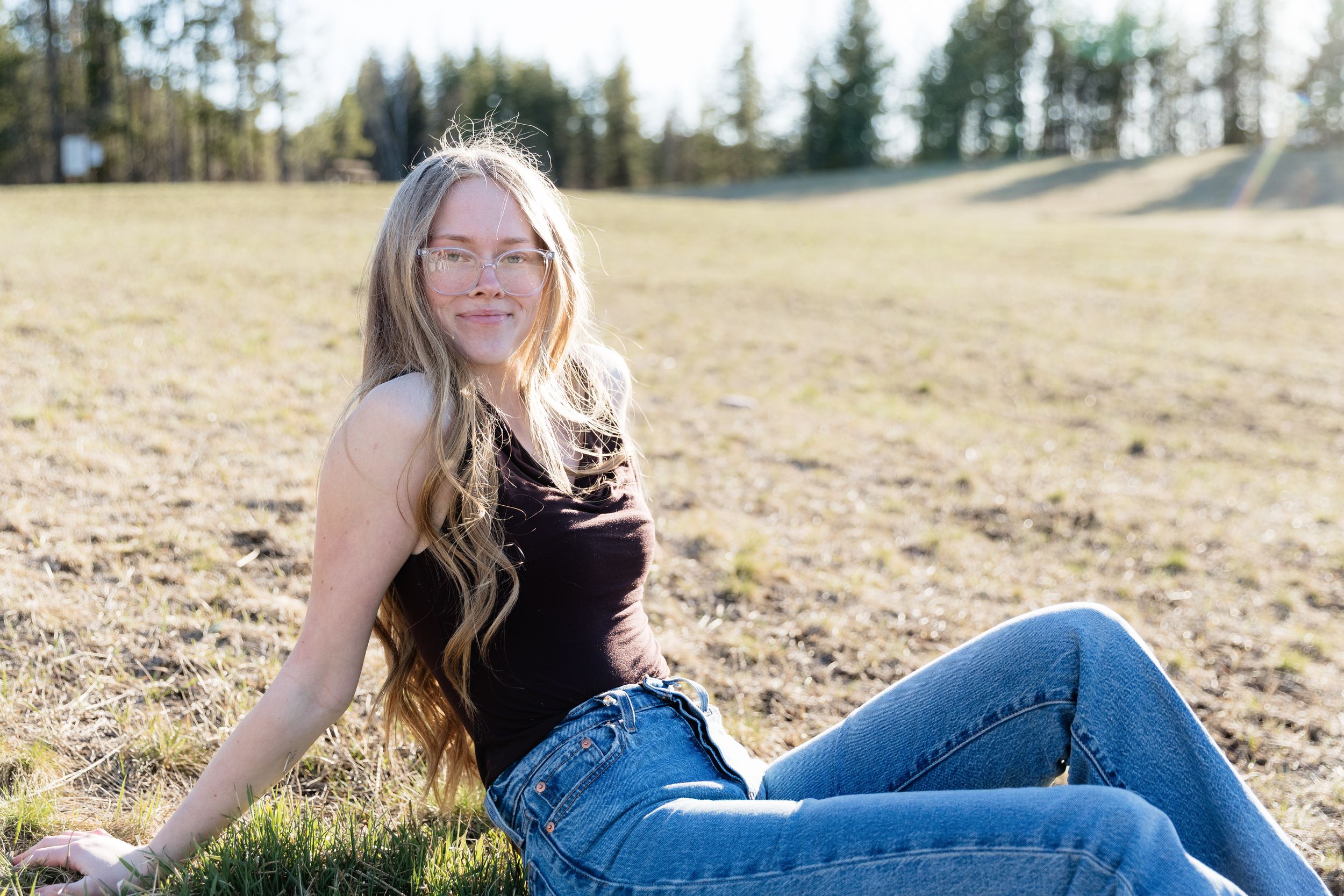 Young woman with long blonde hair sitting in a sunny field, wearing jeans and a dark top, smiling outdoors.