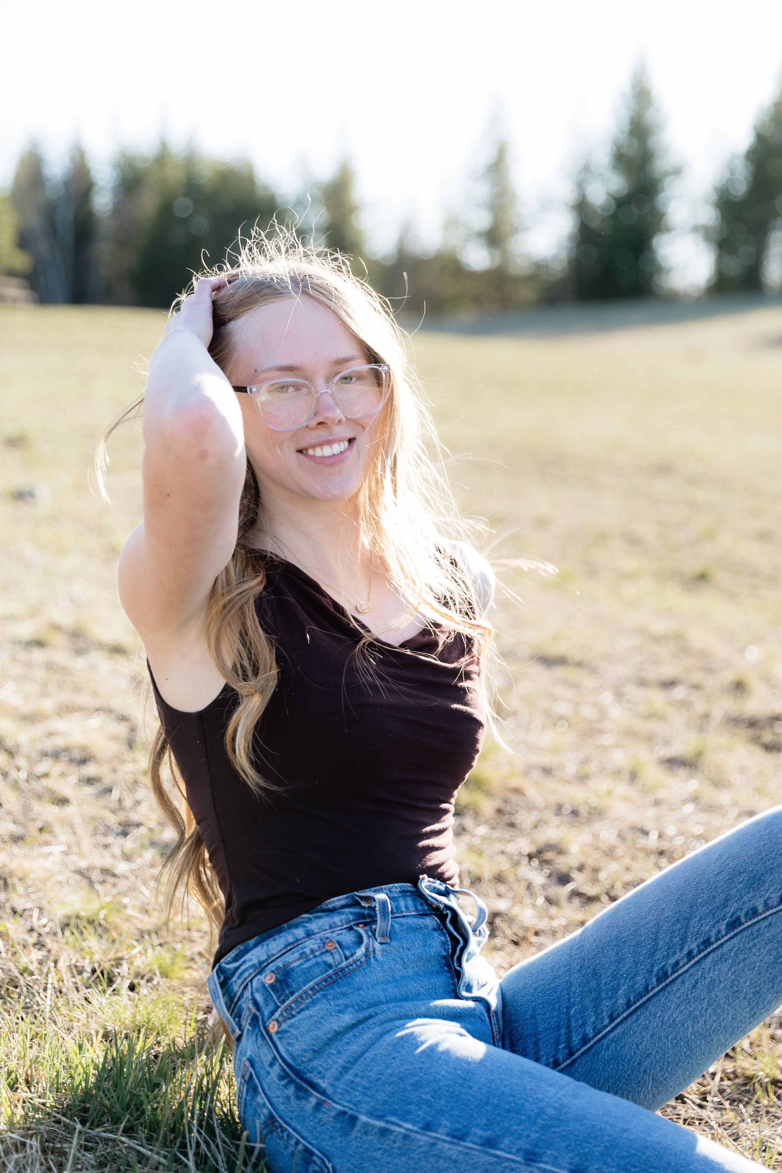 Smiling young woman with long blonde hair and glasses sitting in a sunny field wearing a black tank top and jeans.