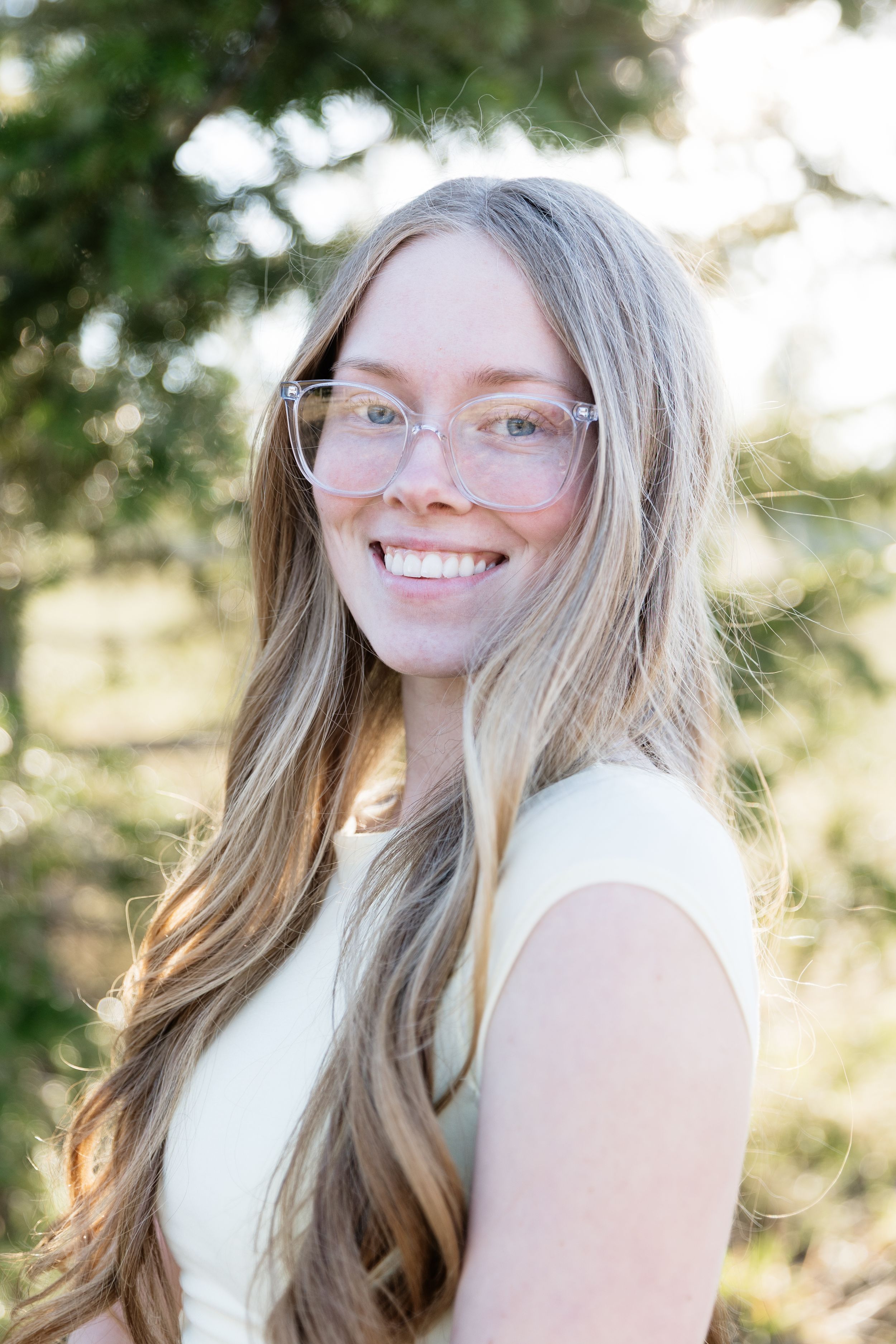 Smiling young woman with long blonde hair and clear glasses outdoors near green trees in natural light.