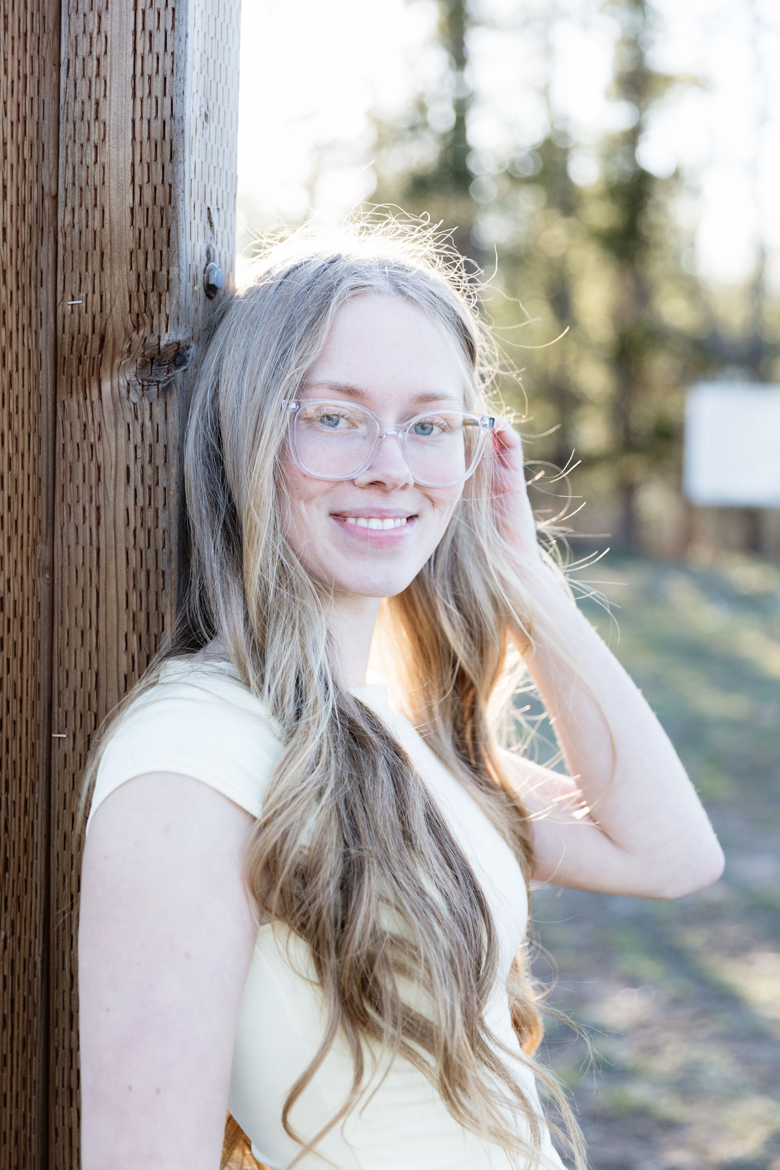 Young woman with long blonde hair and glasses smiling outdoors near a wooden post on a sunny day.