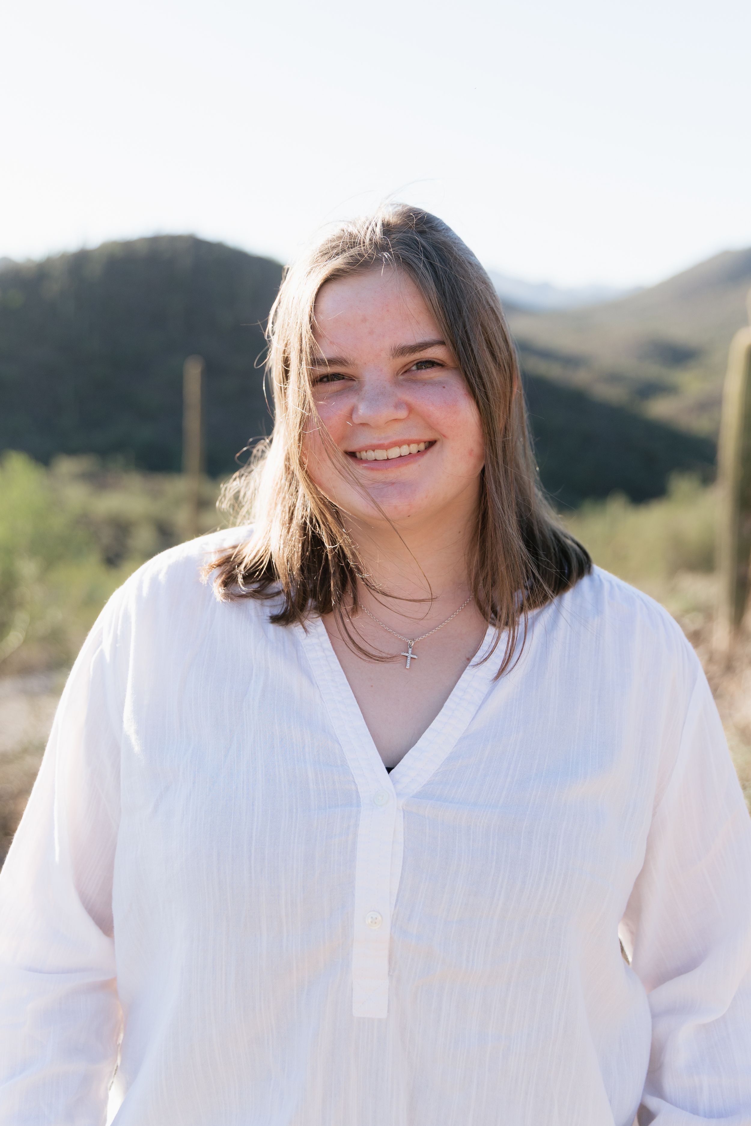 Young woman smiling outdoors in white linen shirt with desert mountains and cacti in background.