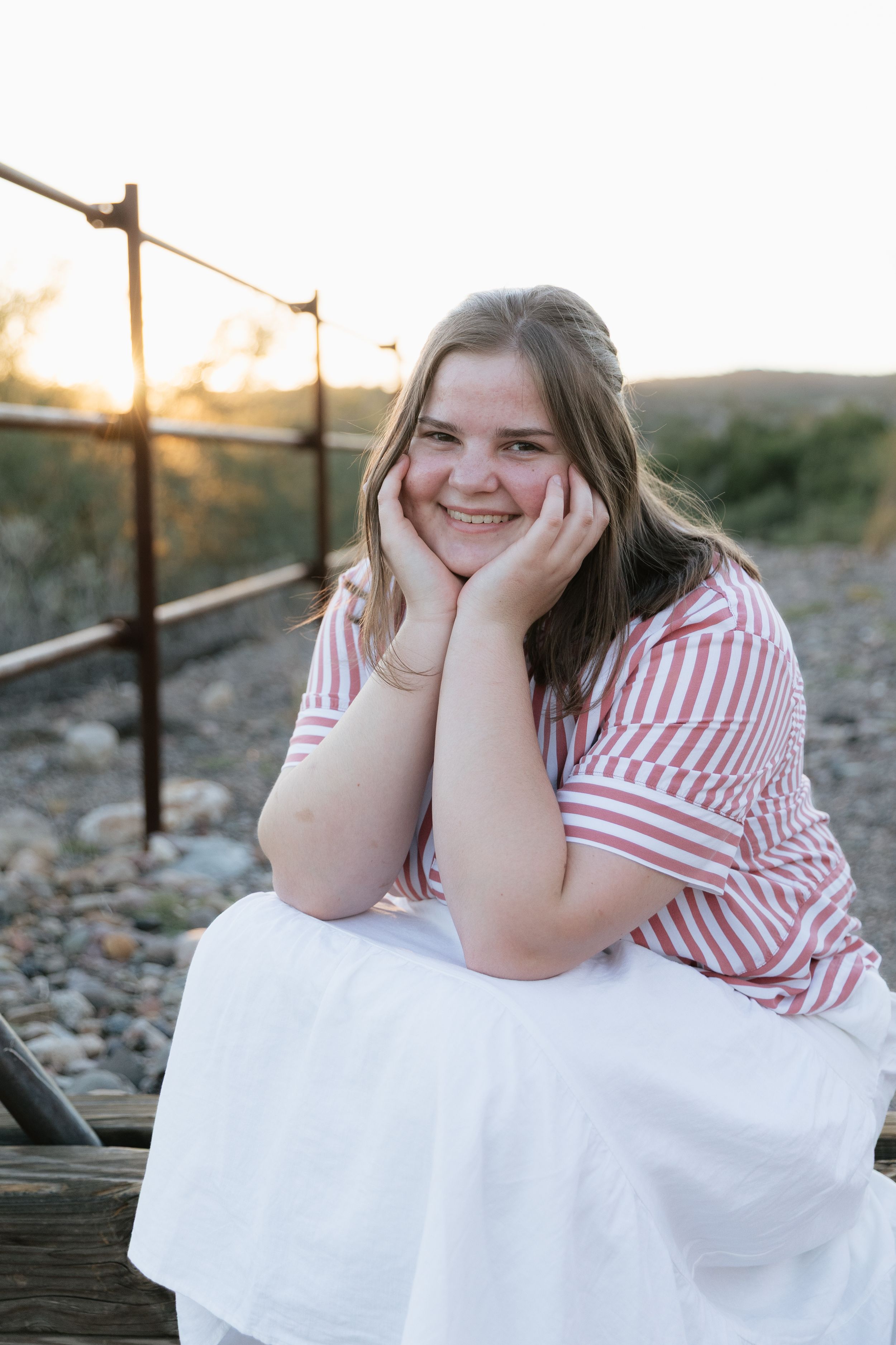 Smiling young woman in red striped top and white skirt sitting outdoors at golden hour sunset.