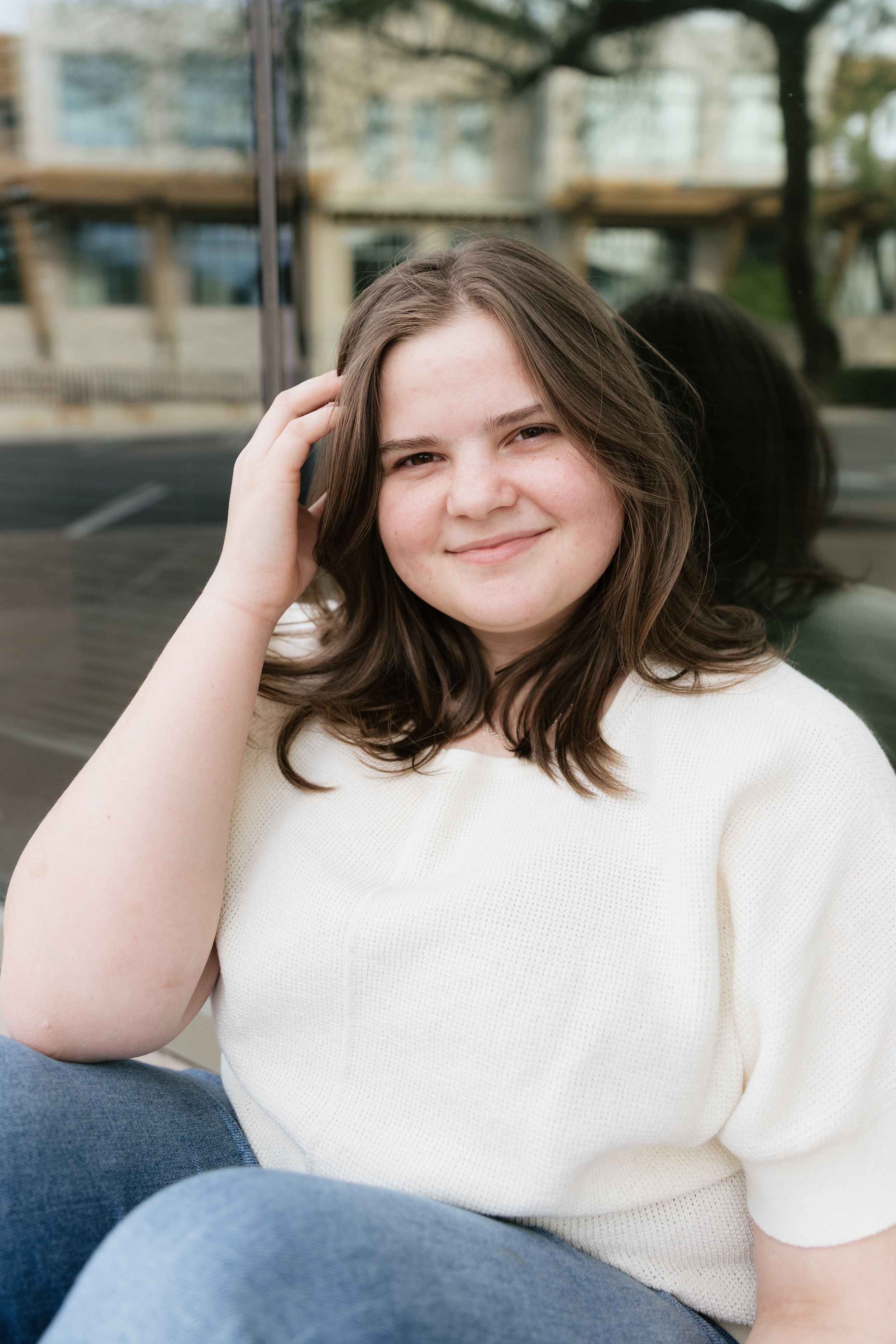 Young woman in cream sweater smiling while sitting outdoors with hand in her brown hair.