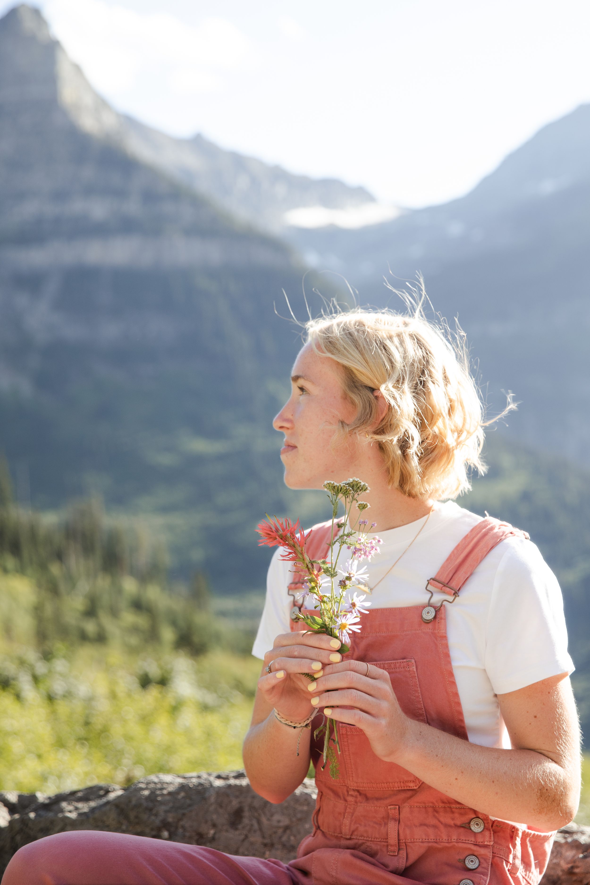 Young blonde woman in pink overalls holding wildflowers, sitting outdoors with scenic mountain backdrop.