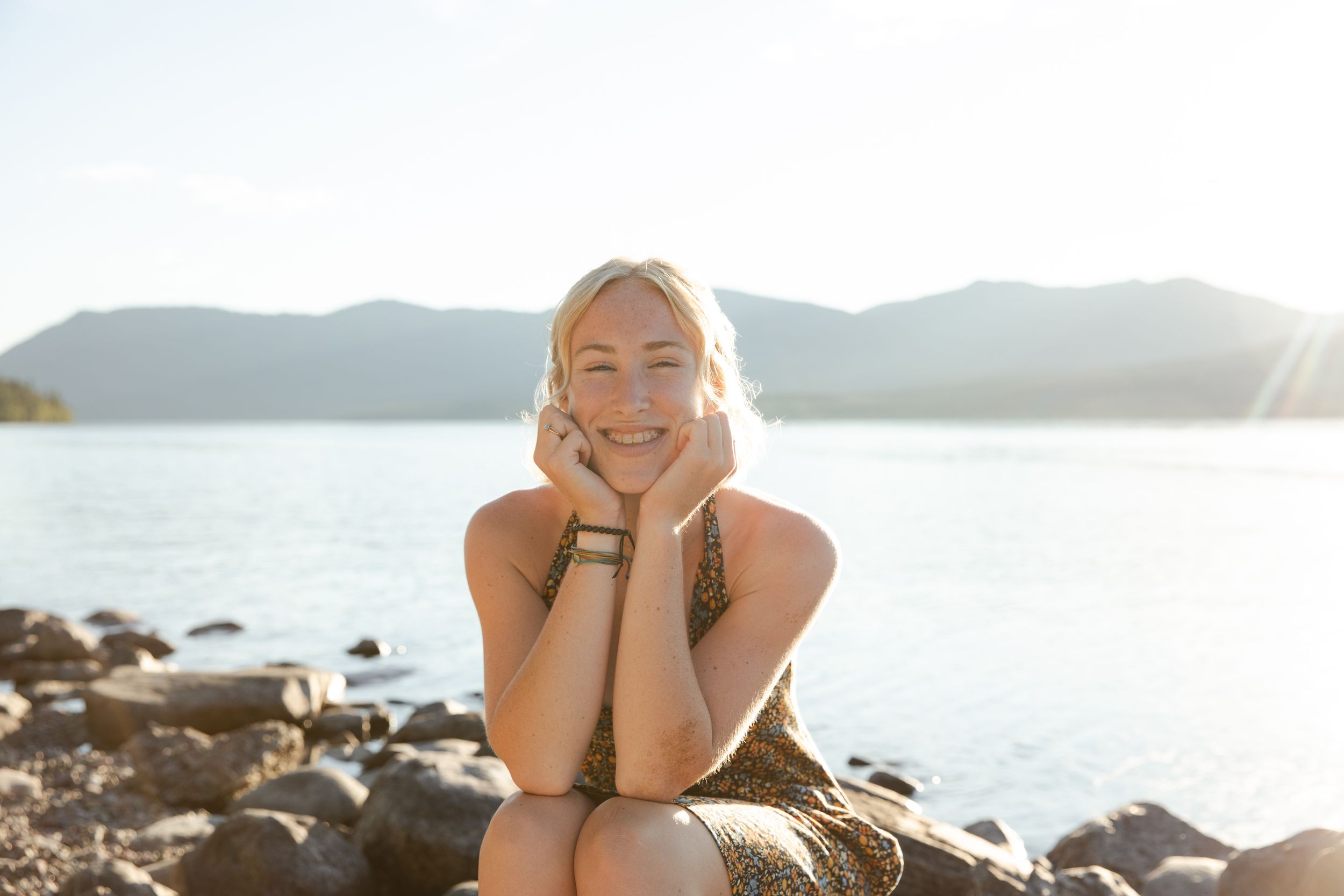 Smiling blonde woman sitting on rocky shore with mountain lake and bright sunlight in background.