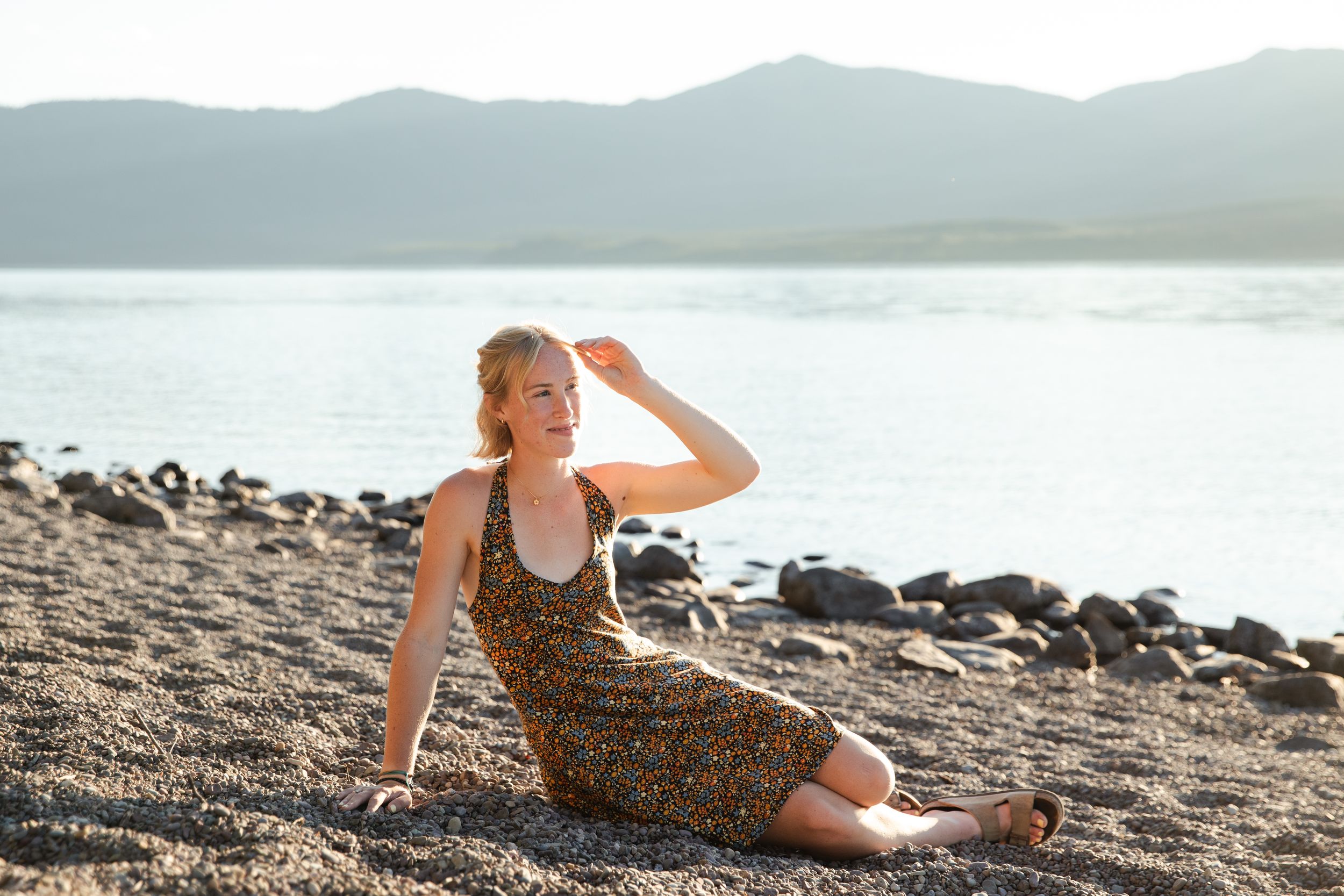 Woman in floral dress sitting on rocky beach shore with mountains and calm water in background during golden hour.