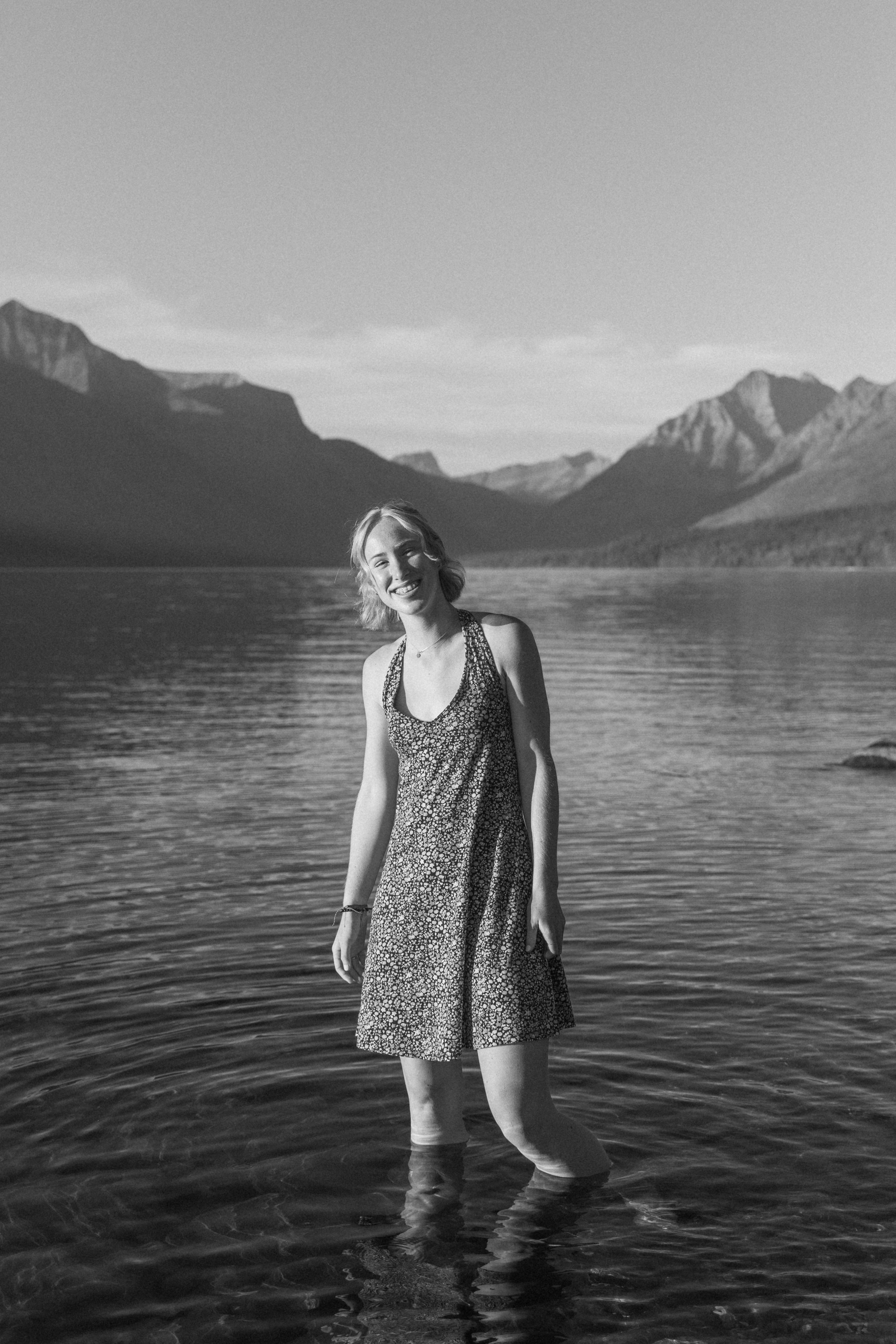 Black and white photo of a woman in a floral dress standing in a mountain lake with scenic peaks in the background.