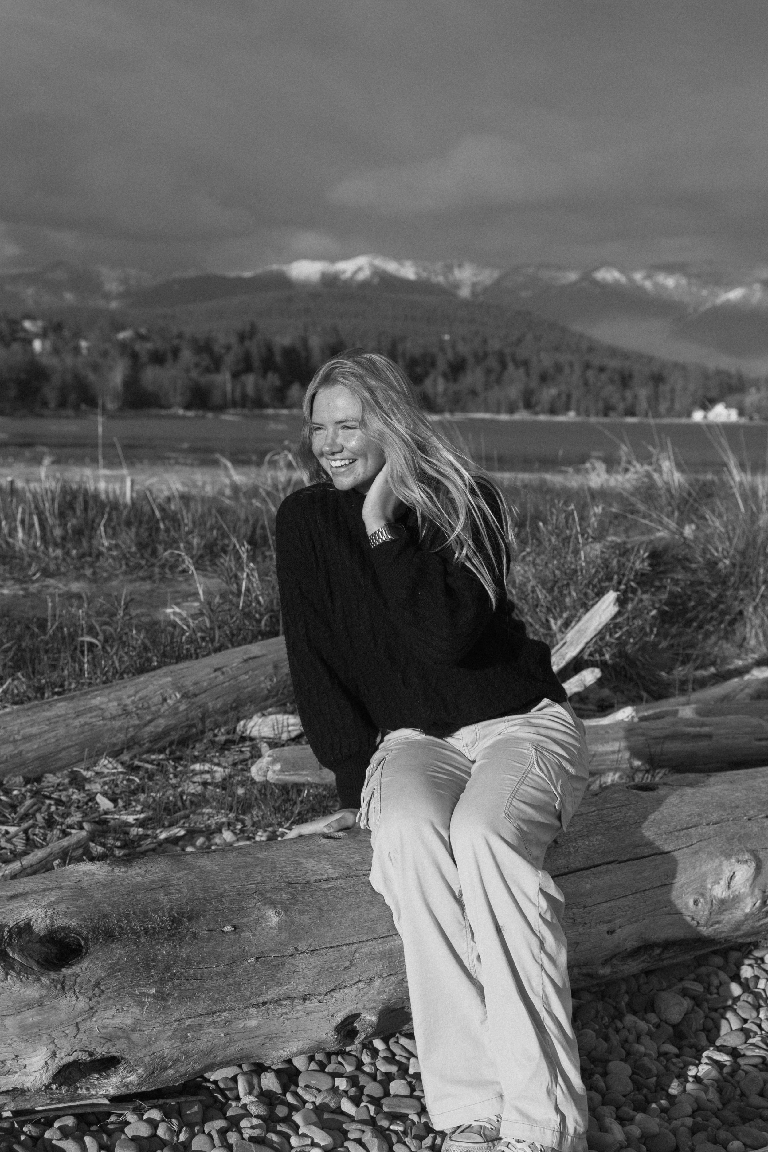 Black and white photo of a laughing woman sitting on driftwood logs with mountains and forest in background.